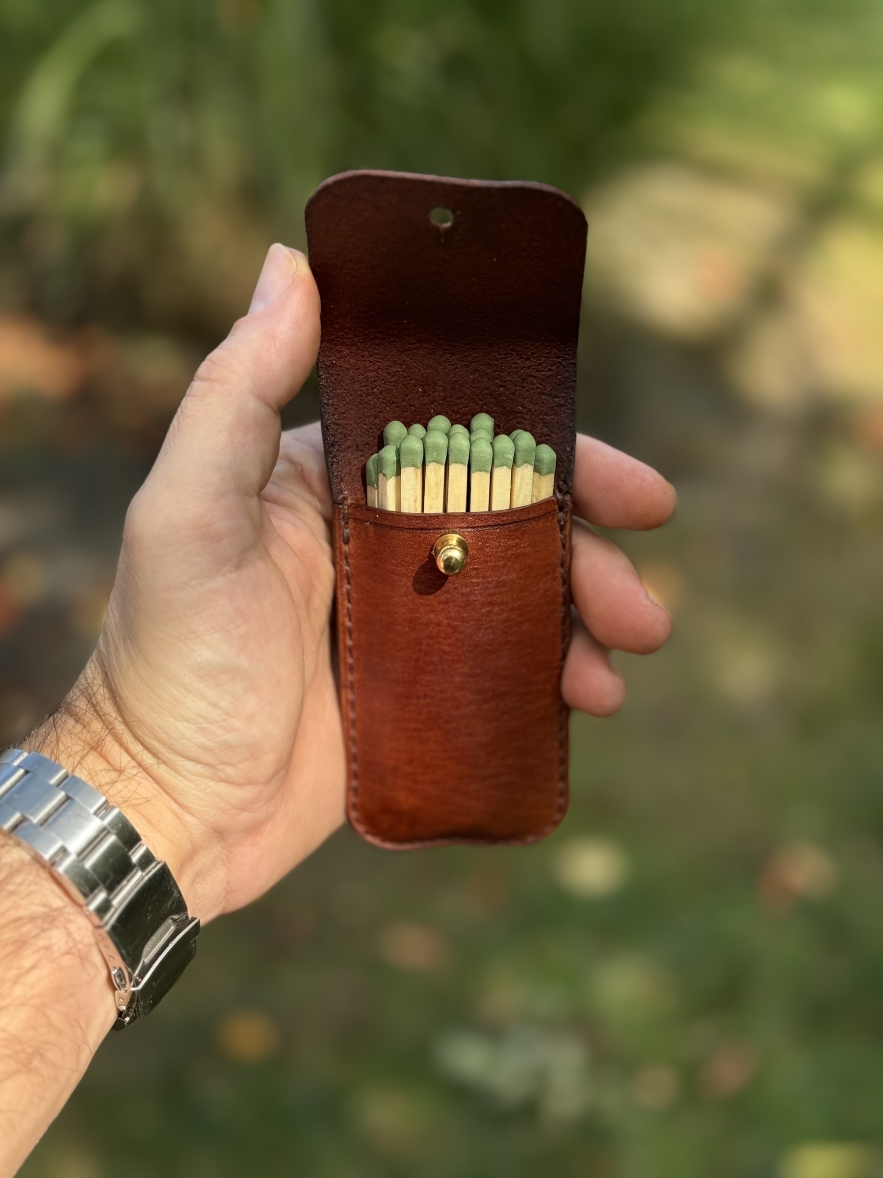 A hand is holding a brown leather match holder containing green-tipped cigar matches.