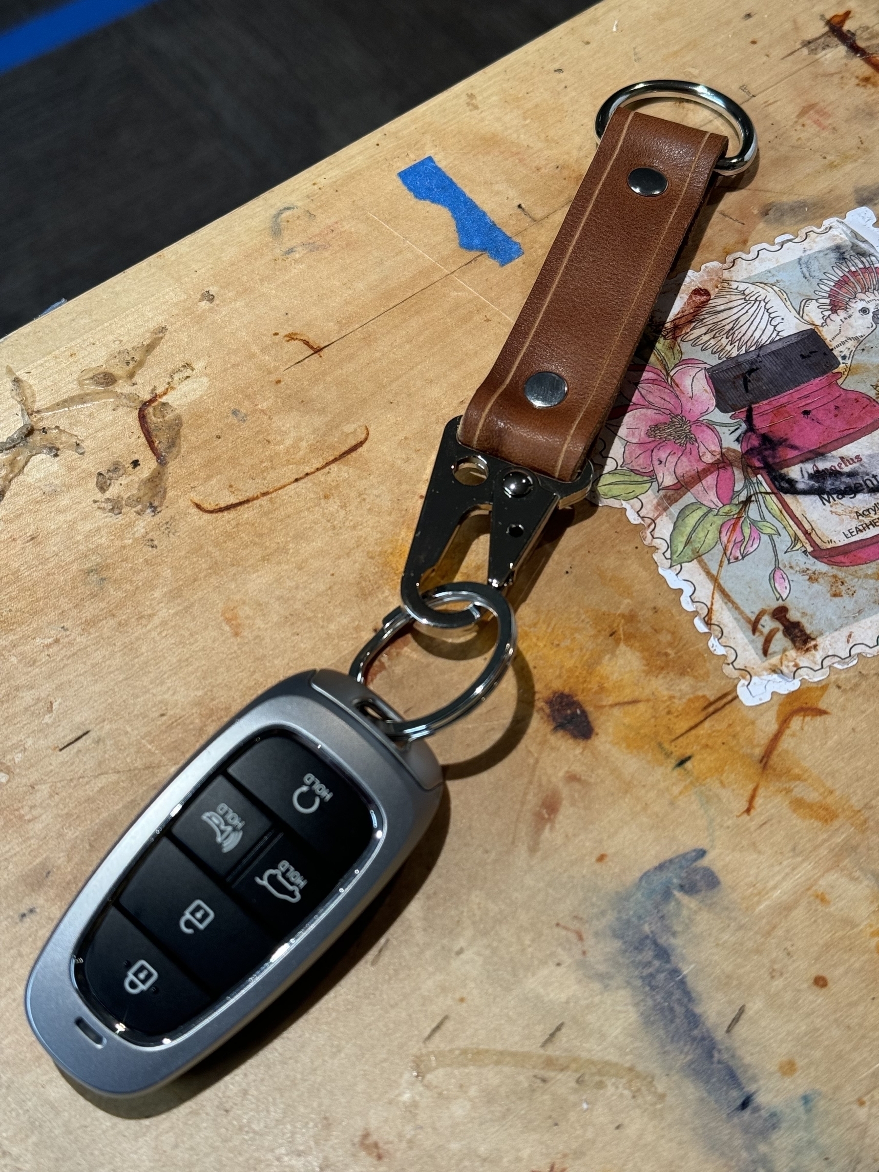 A car key fob attached to a leather keychain is resting on a wooden surface with a decorative sticker nearby.