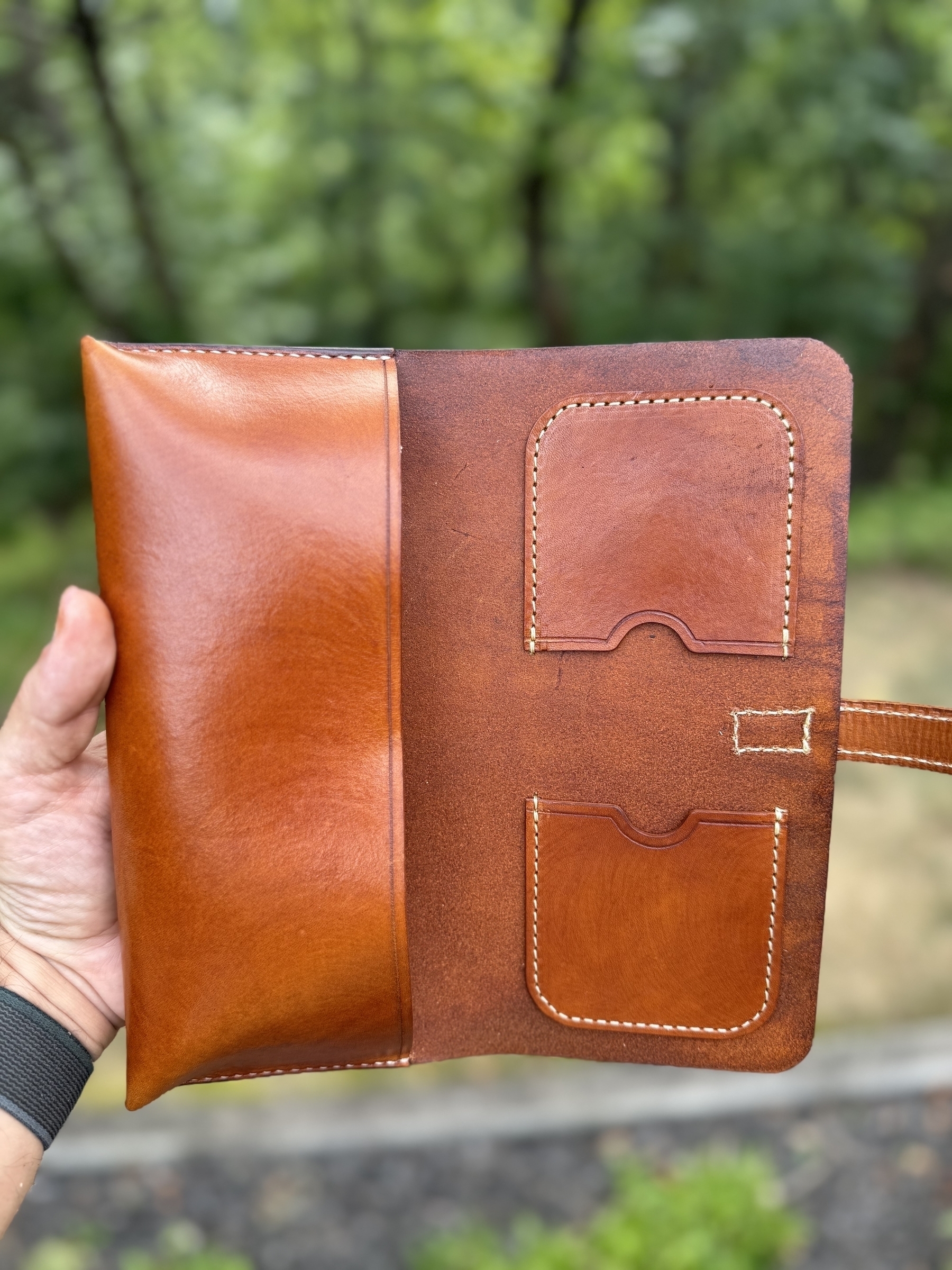 A hand is holding an open brown leather cigar case with multiple compartments, in an outdoor setting with greenery in the background.