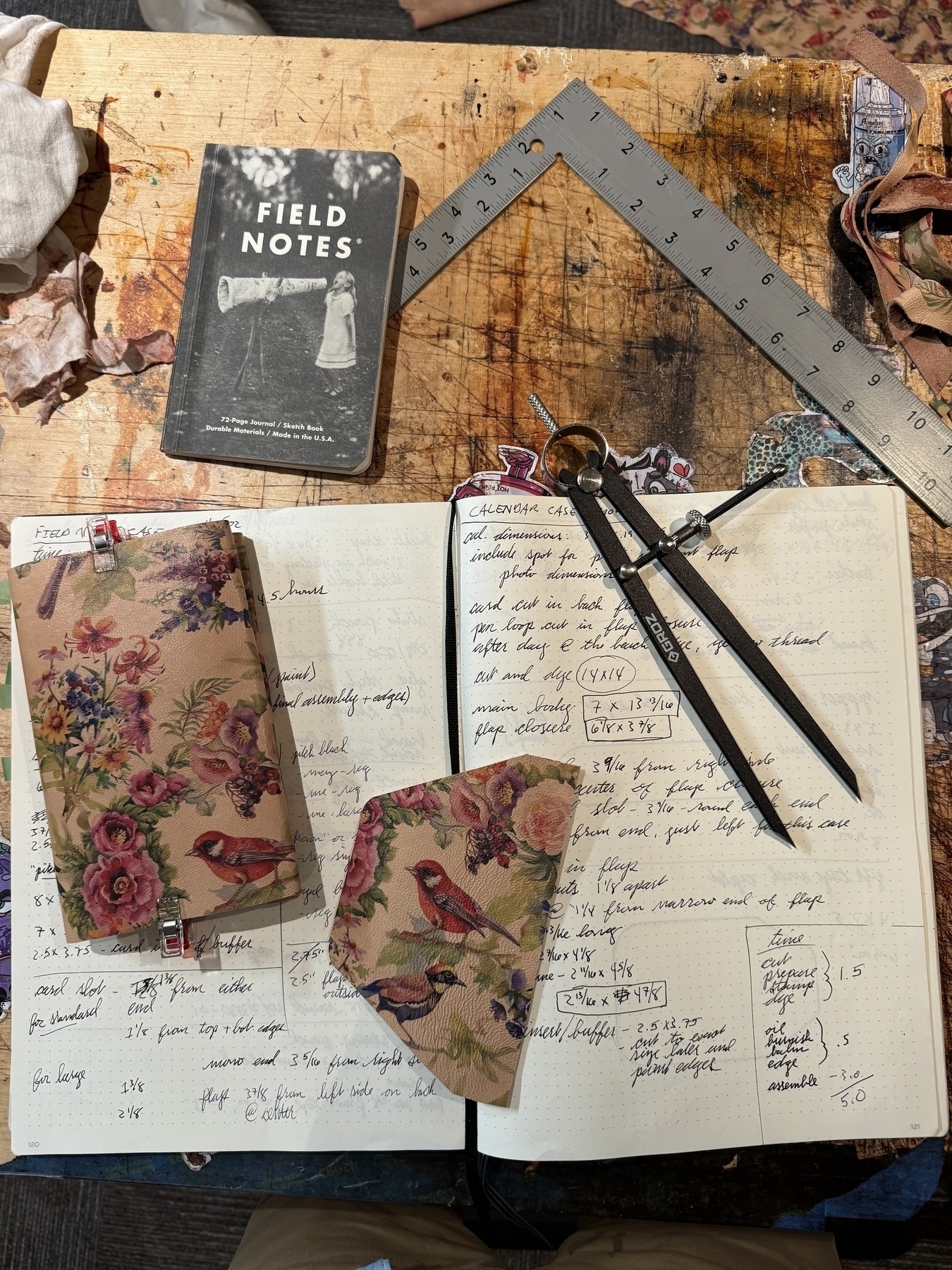 A workbench is cluttered with a patterned notebook, fabric with floral and bird designs, an open lined journal with handwritten notes, a black "Field Notes" booklet, a ruler, and drafting tools.