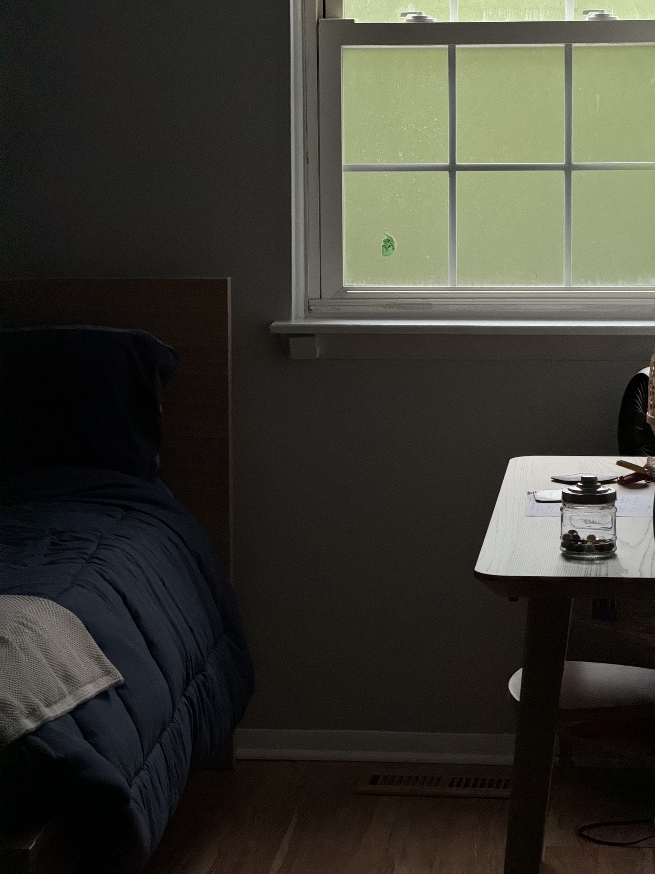 A dimly lit room features a bed with dark bedding on the left, a rain-covered window with white trim in the center, and a table with items on it to the right.