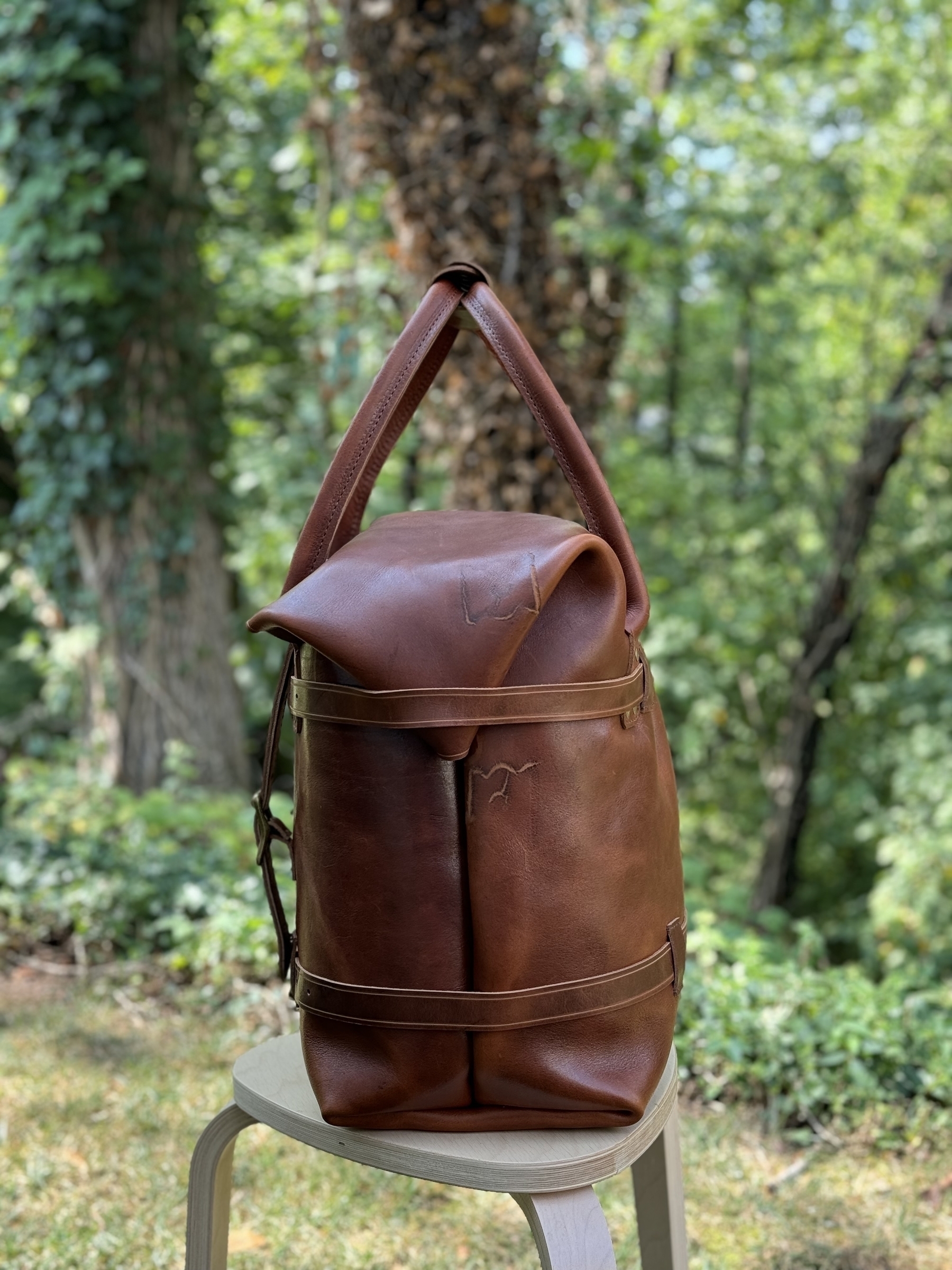 A brown leather weekend bag with a rolled top sits on a small light-colored stool in a lush, green outdoor setting.