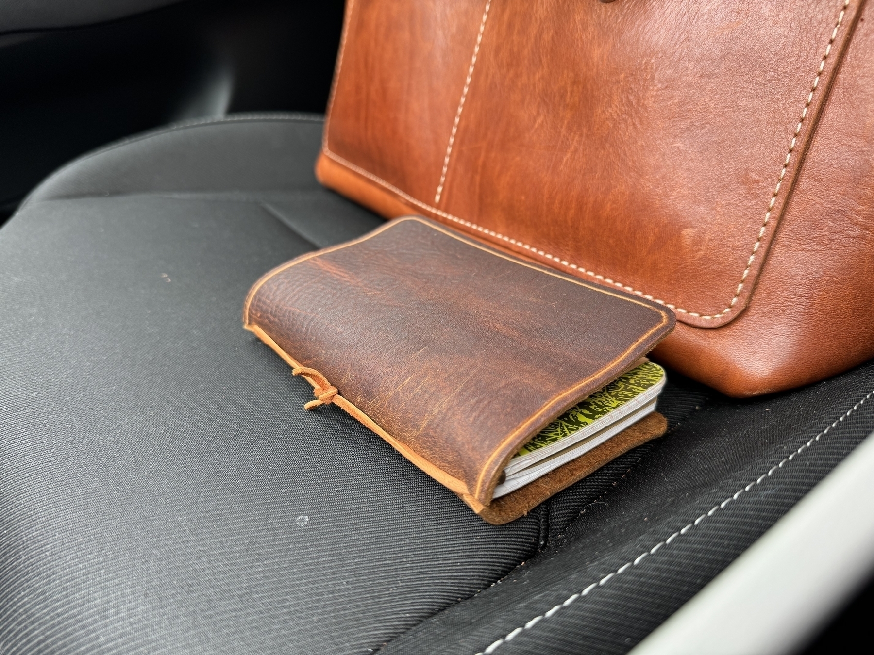 A leather journal rests on the seat of a car next to a brown leather bag.