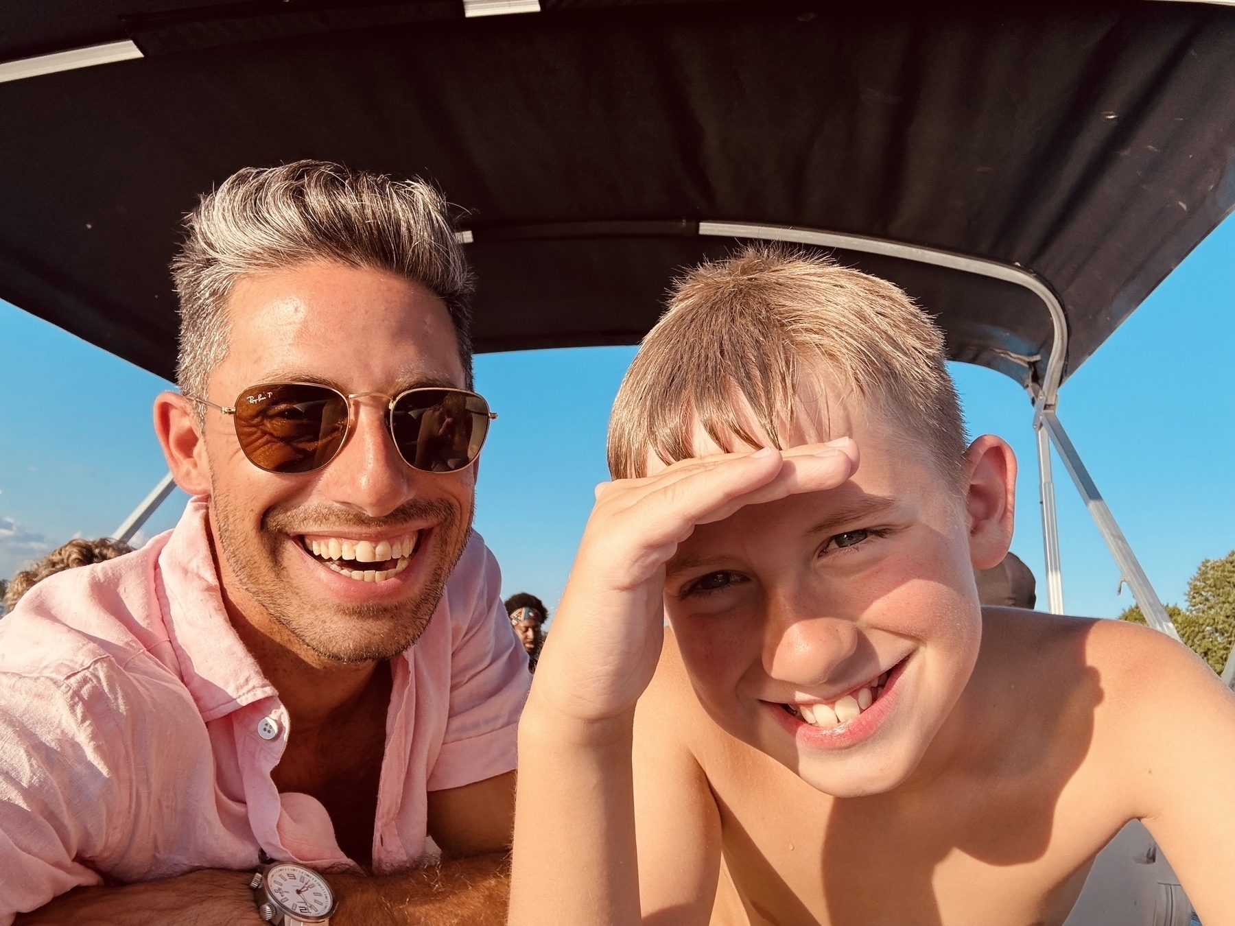 A man (Aaron) and a child (Mozzie), both smiling and appearing happy, are enjoying a sunny day under a canopy of a pontoon boat.