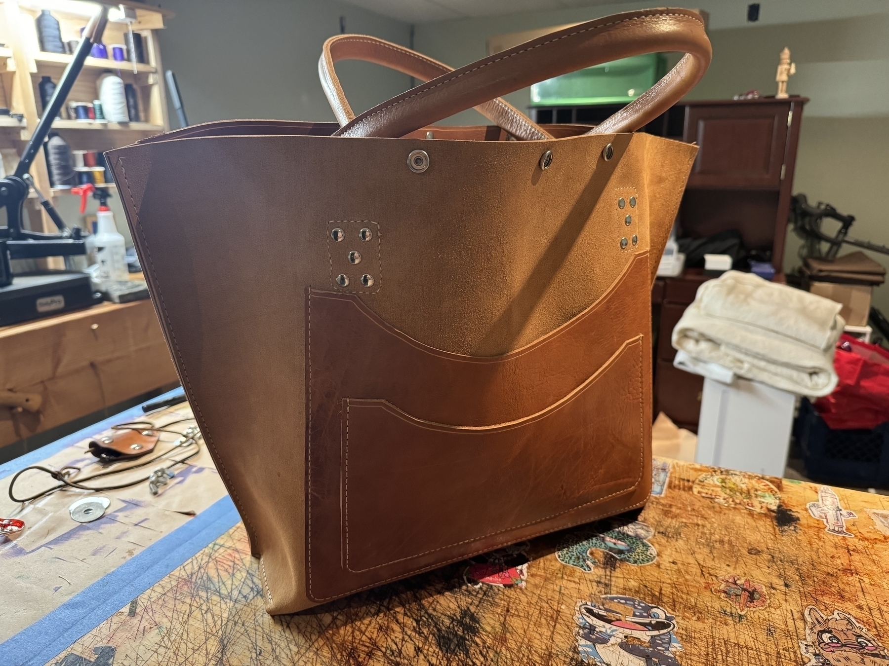 A brown leather handbag with visible stitching and a front pocket is displayed on a cluttered table in what appears to be a workshop.