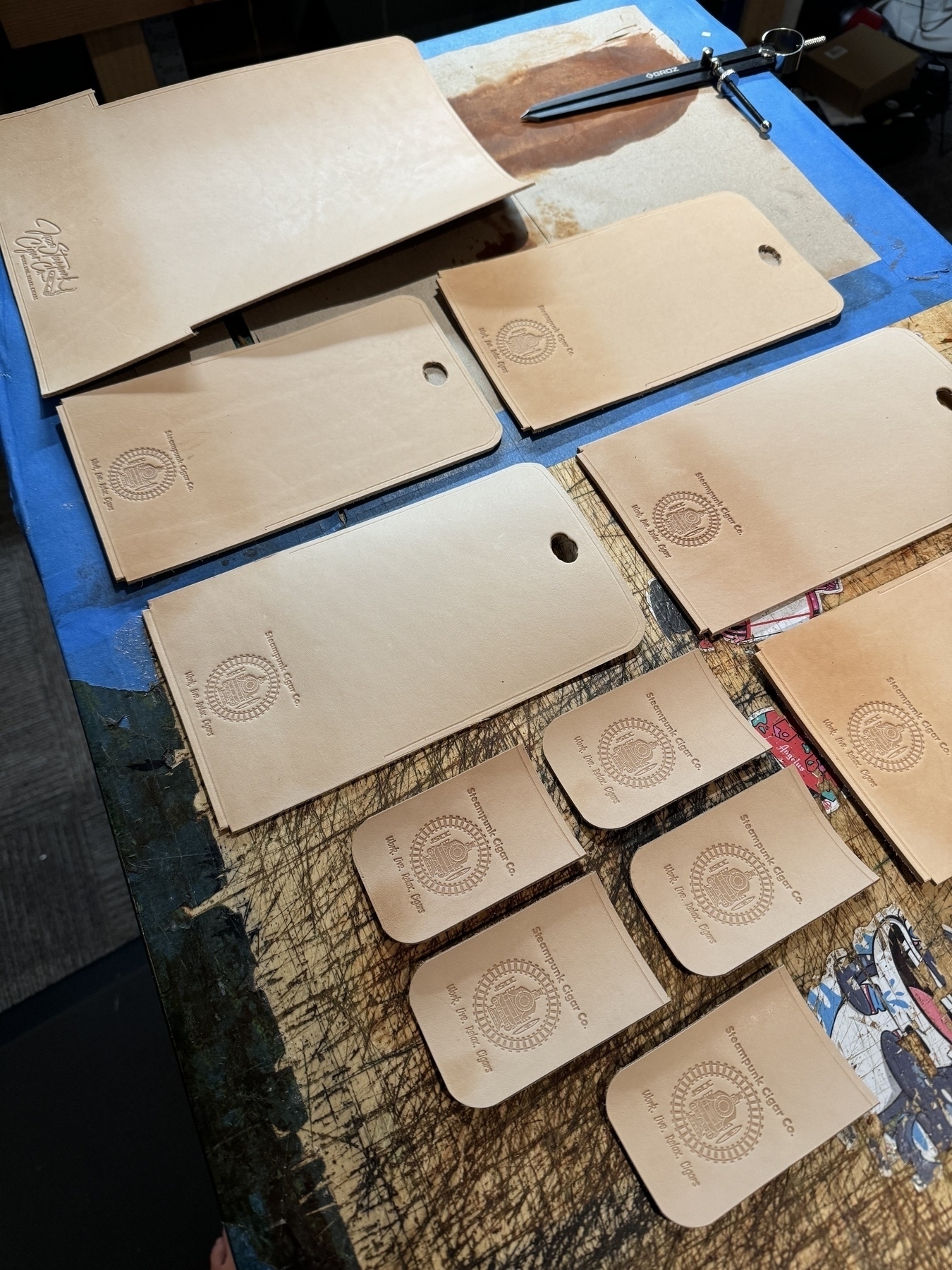 A collection of variously sized brown leather tags, each embossed with a logo and text, are arranged on a table alongside a pen and some leatherworking tools.