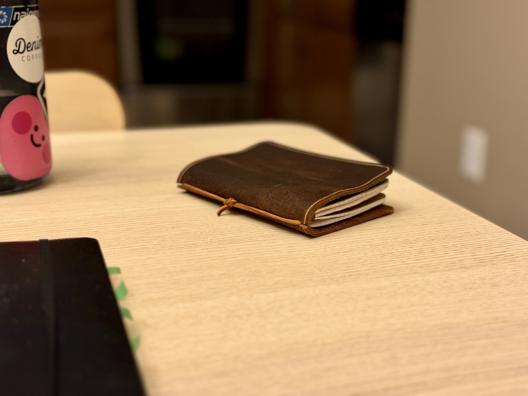 A leather-bound book and a bottle with a pink sticker on it are placed on a light-colored wooden table.