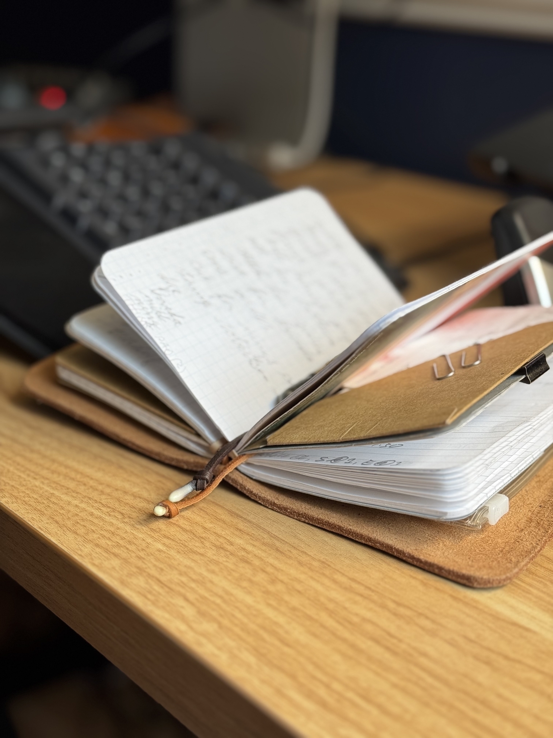 A notebook filled with handwritten notes and a pen resting on top sits on a wooden desk beside a keyboard.