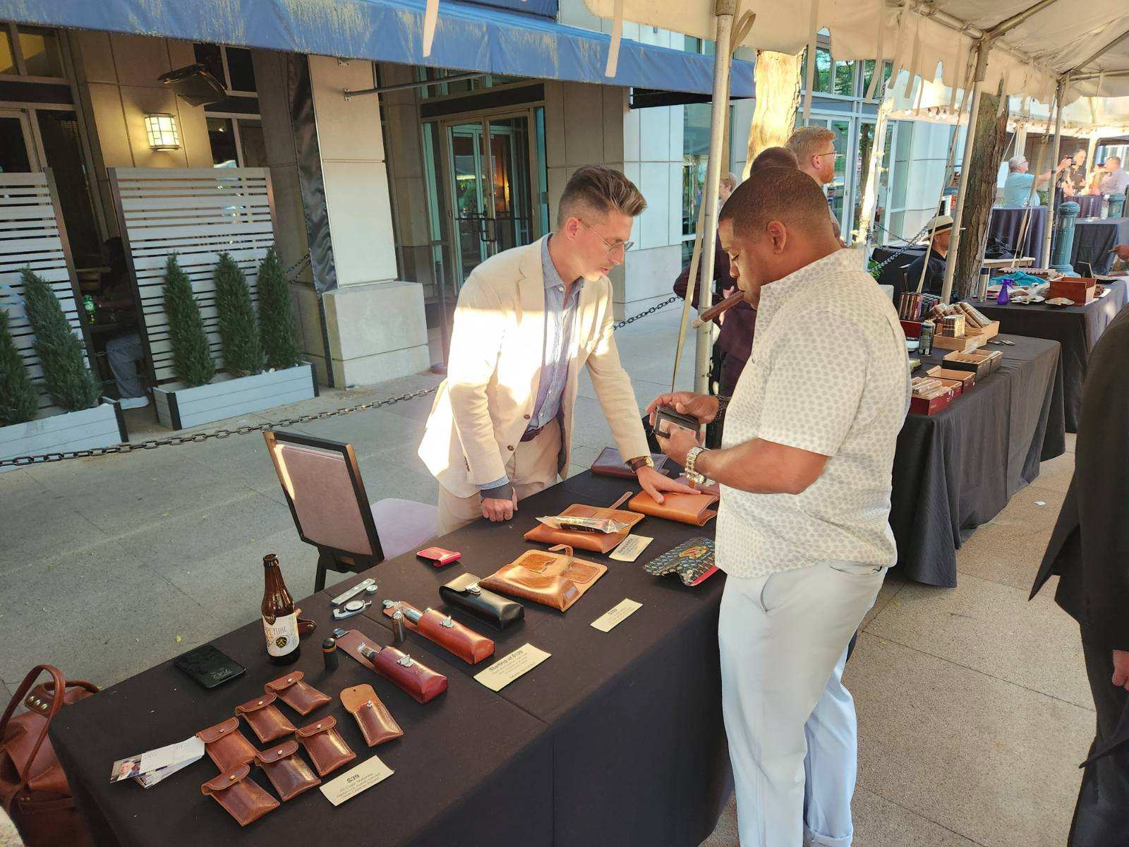 A vendor at an outdoor market stall is showing leather goods to a customer.