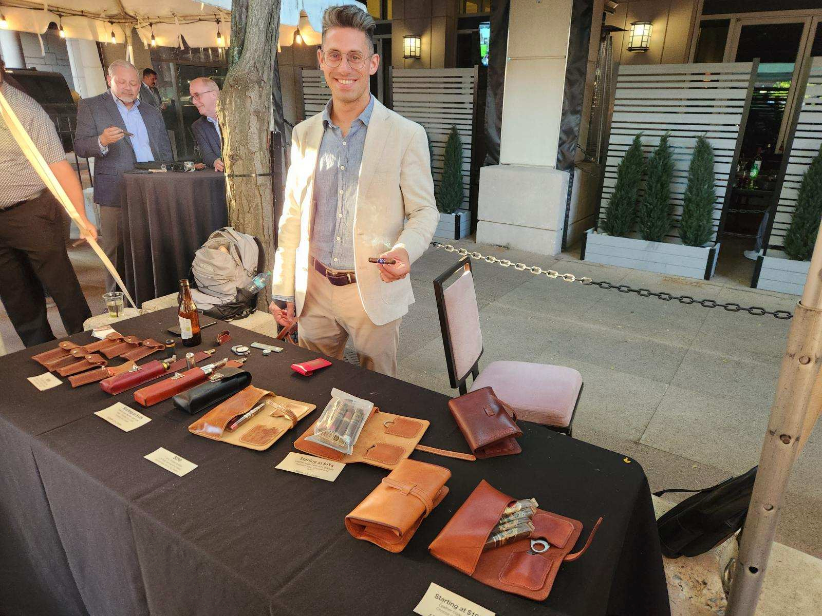 A man is standing behind a table displaying various leather goods at an outdoor event.