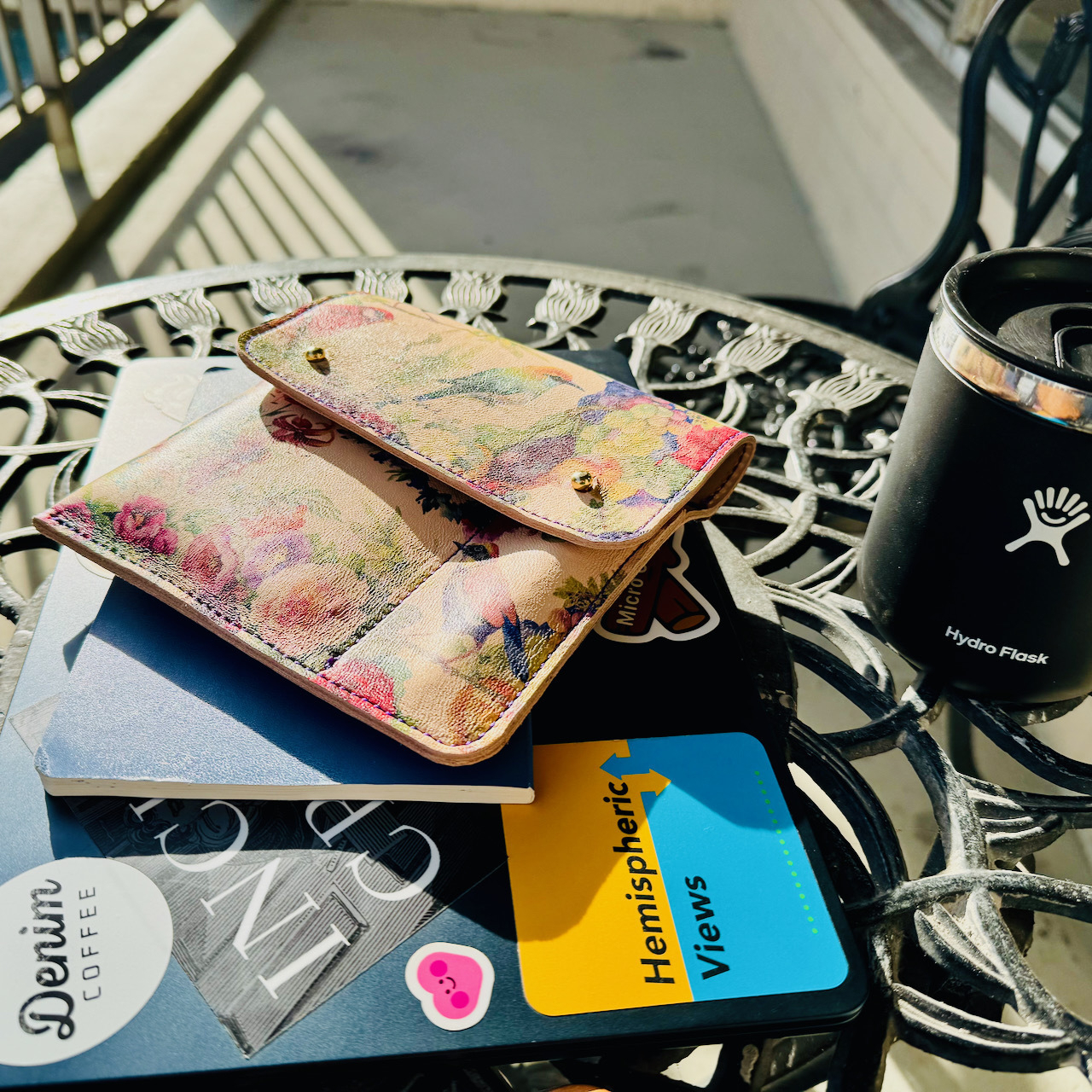 laptop adorned with stickers, notebook, and a beautiful leather pouch stacked on a table outisde in the sun