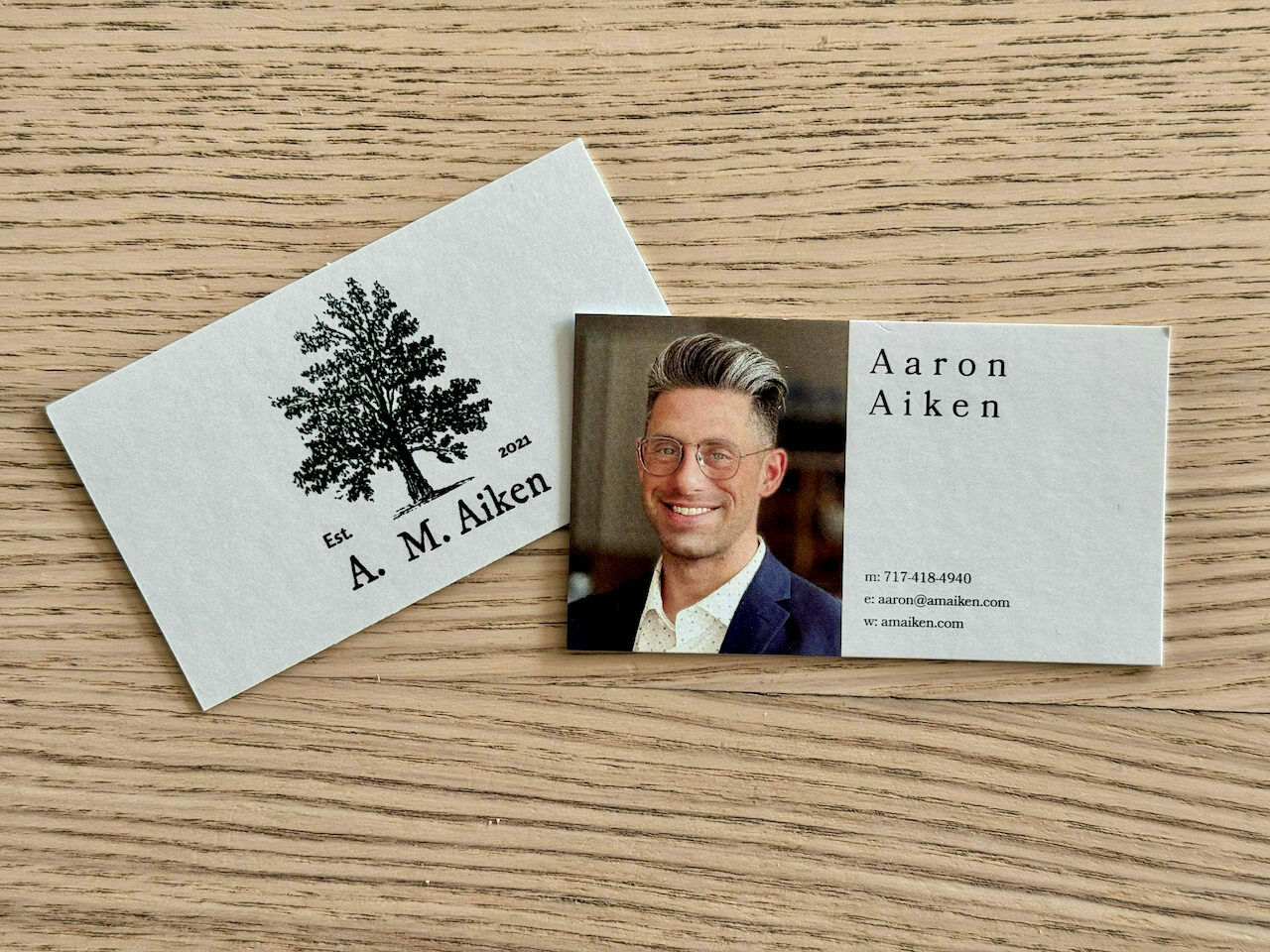 new business cards for A. M. Aiken - shown on a woodgrain table, one card showing the front of the card which has a nice whimsical tree on it, the other card showing the back which has a nice photo of Aaron and his contact details.