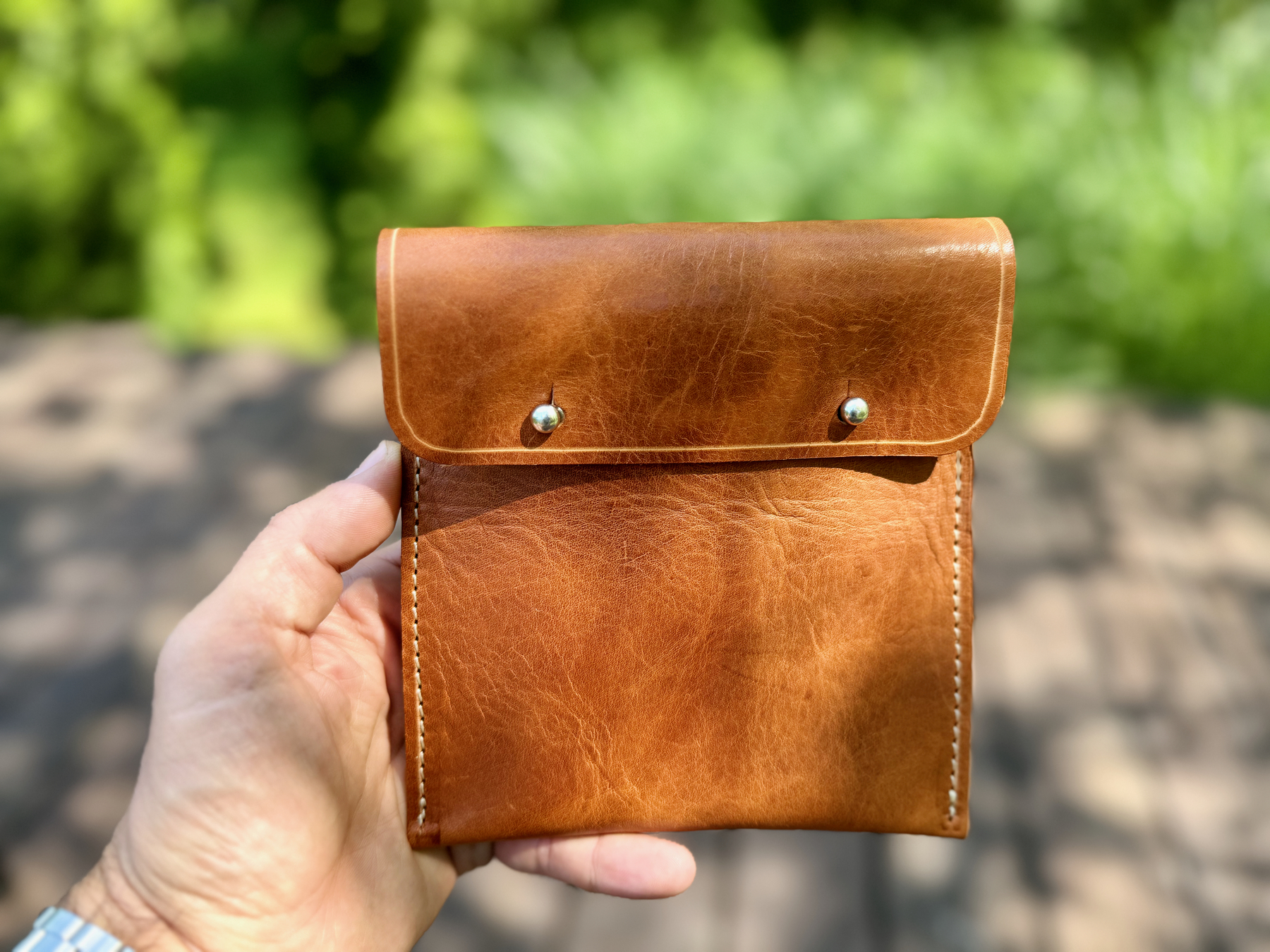 A person's hand holds a brown leather pouch with two metal snap buttons, against a blurred outdoor background.