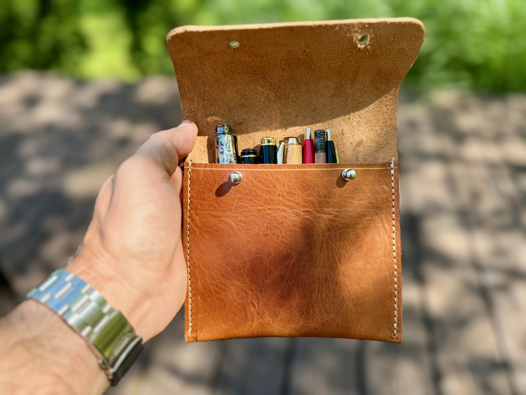 A hand holds an open leather pouch containing multiple pens and a pencil.