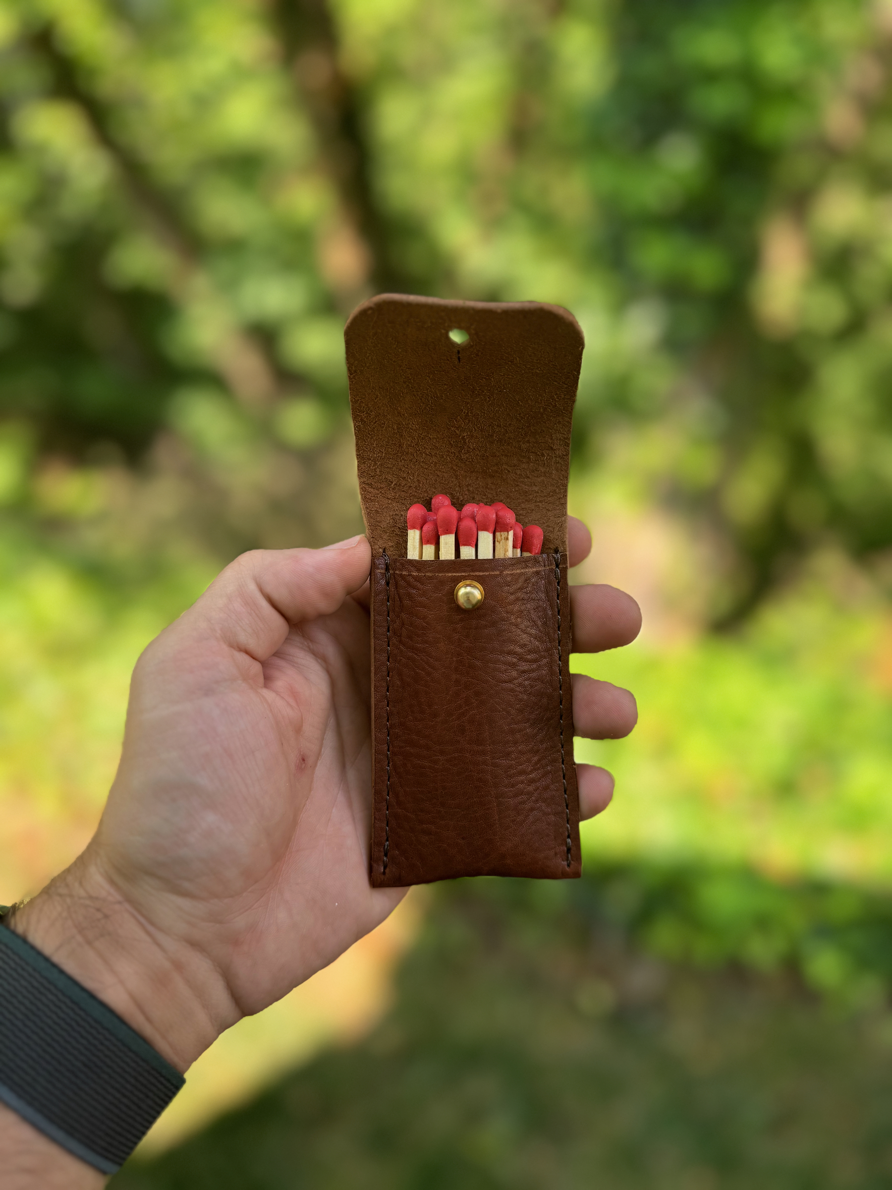 A person is holding an open brown leather pouch containing matches, with a green, outdoor background.