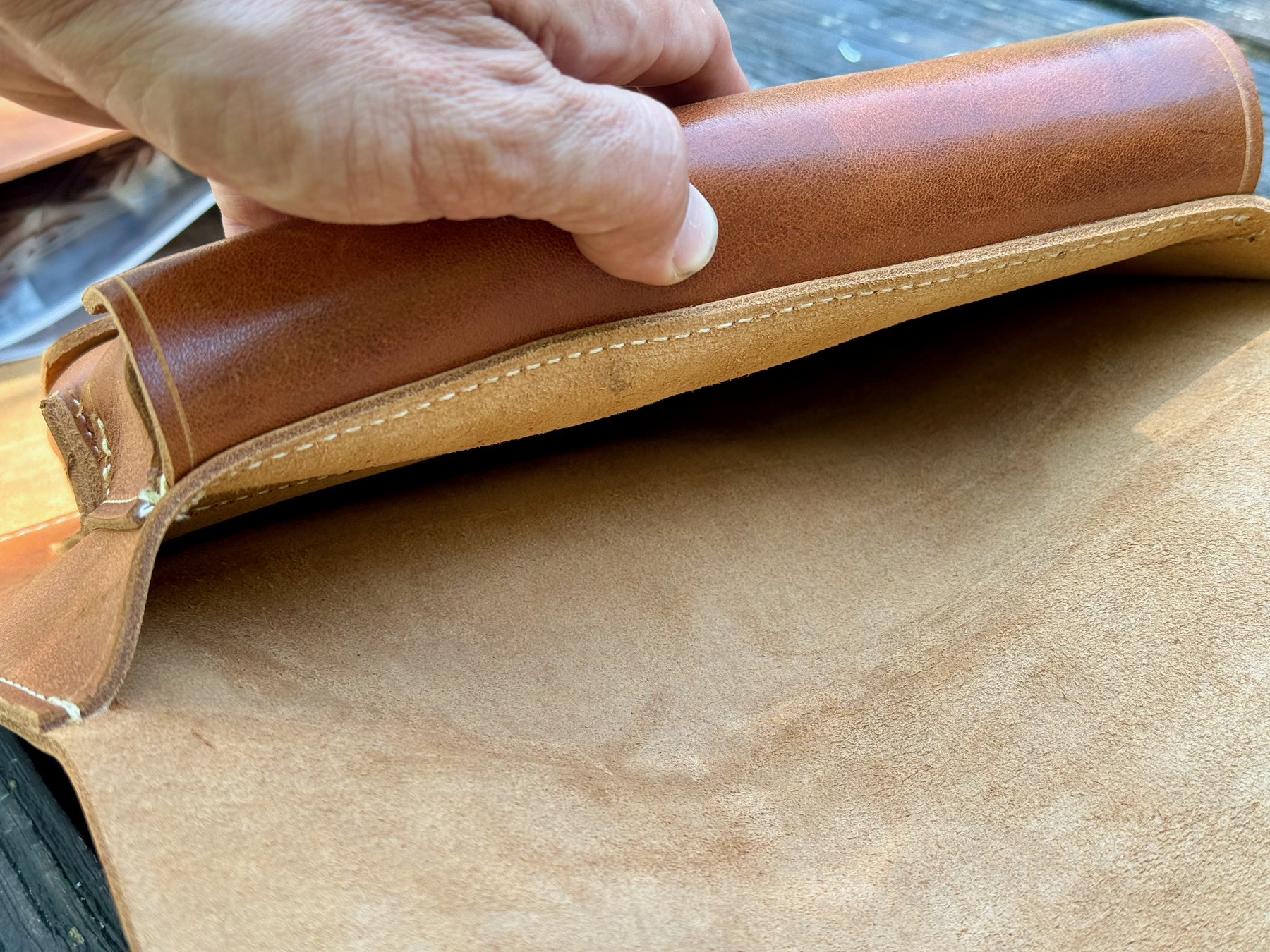 A hand is examining the inside of a partially open, tan-brown leather cigar case, against a wooden background.