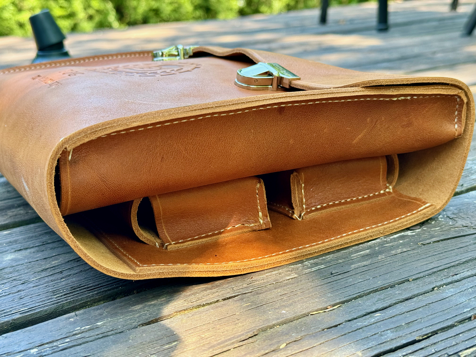 A close-up of a tan leather cigar case showing its open pocket and detailed stitching, placed on a wooden surface with a blurred outdoor background.