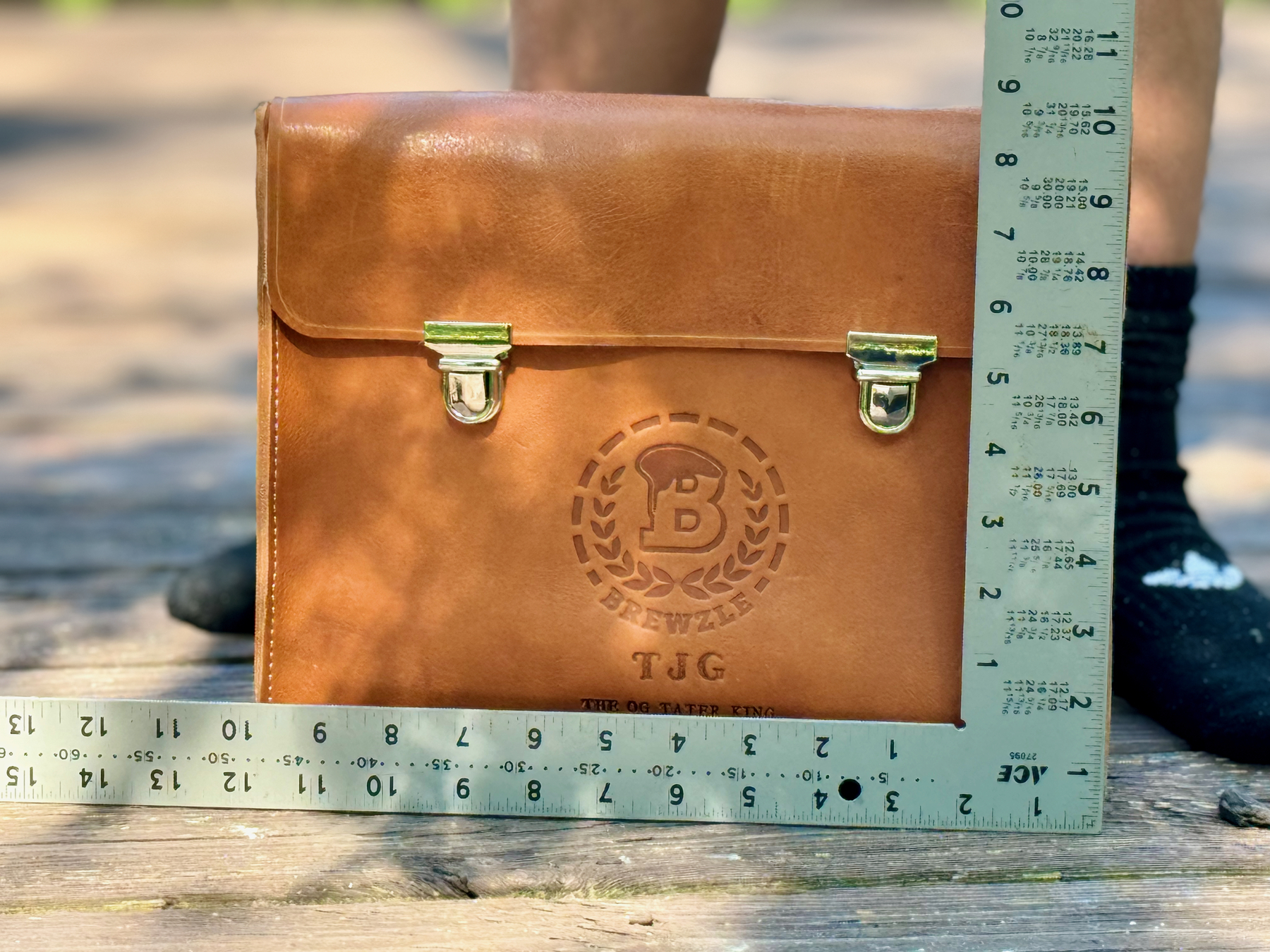 A brown leather cigar case with metal clasps, featuring embossed initials and logo, is being measured with a ruler on a wooden surface.