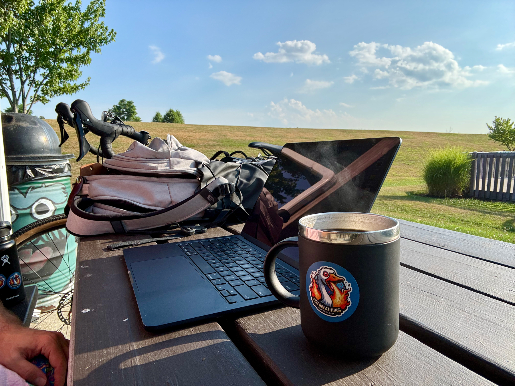 Auto-generated description: A laptop, mug, backpack sit on a picnic table outdoors with a scenic, grassy landscape and trees, and a bicylce in the background.