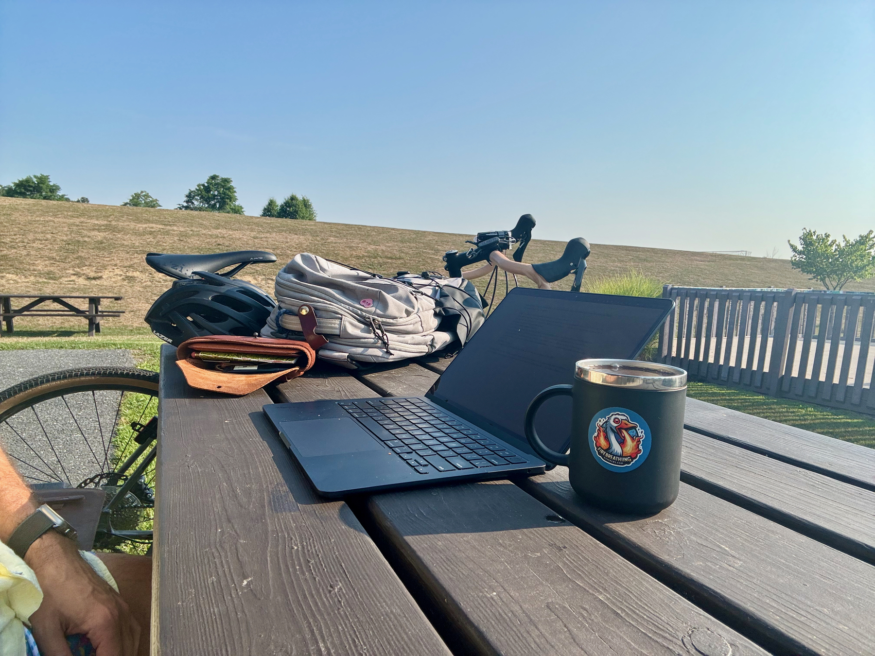 A wooden picnic table holds a laptop, a black mug, a bicycle helmet, a backpack, and a wallet, with a bicycle in the background near an outdoor path under a clear blue sky.