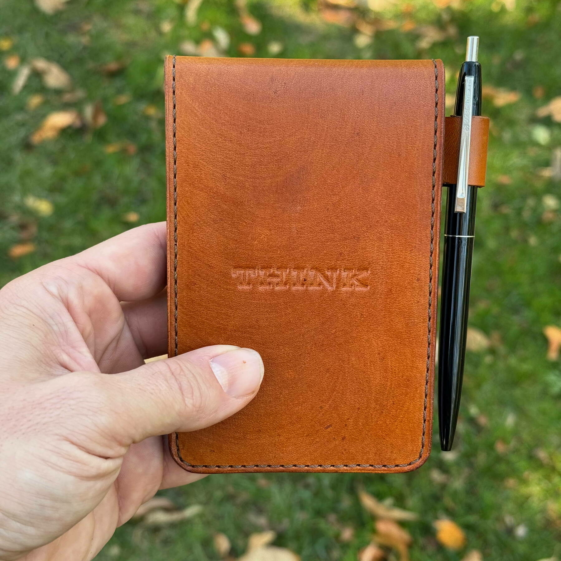 Auto-generated description: A hand is holding a leather notebook with a pen attached on the right side and the word TEFLUNES embossed on the cover, against a grass background.