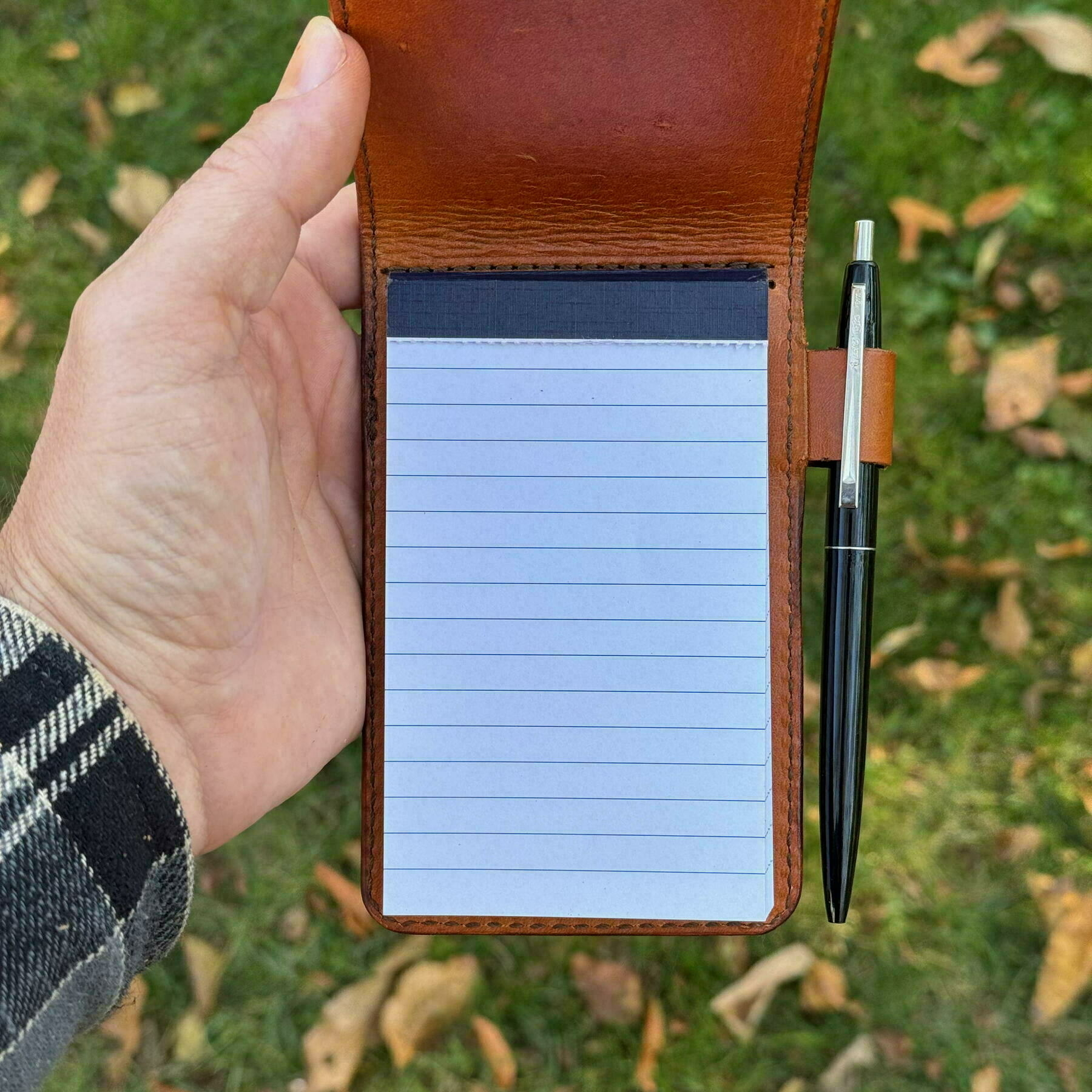 A person is holding an open leather notebook with a pen attached, over a grassy background with fallen leaves.