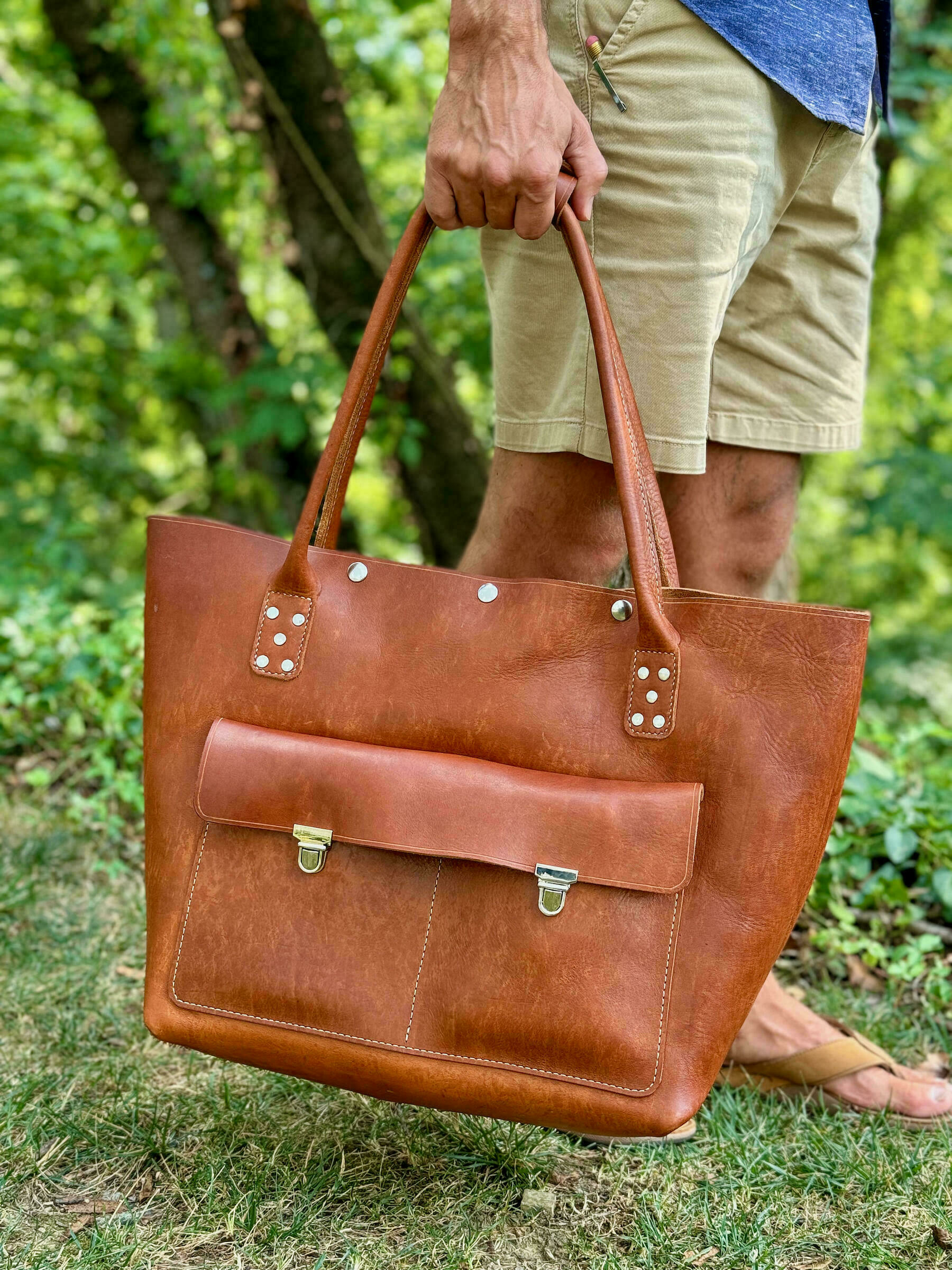 A person is holding a large brown leather tote bag outdoors, with trees and greenery in the background.