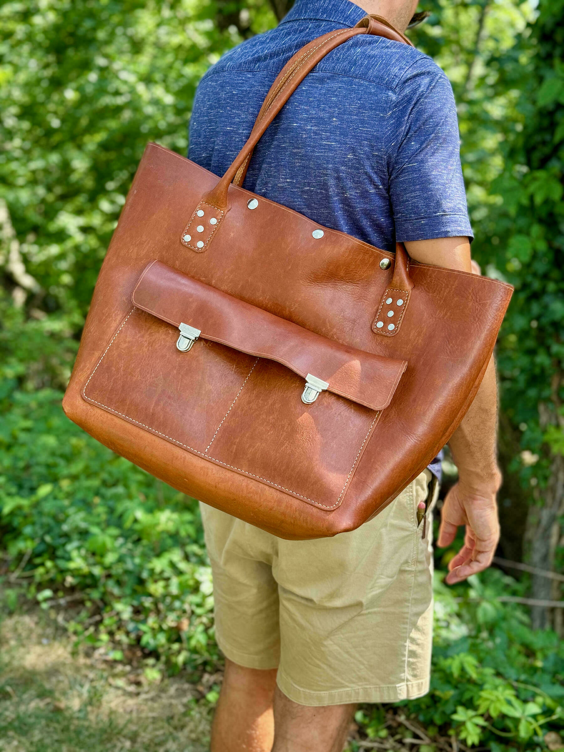 A person is carrying a large brown leather bag over their shoulder while standing outdoors by some greenery.