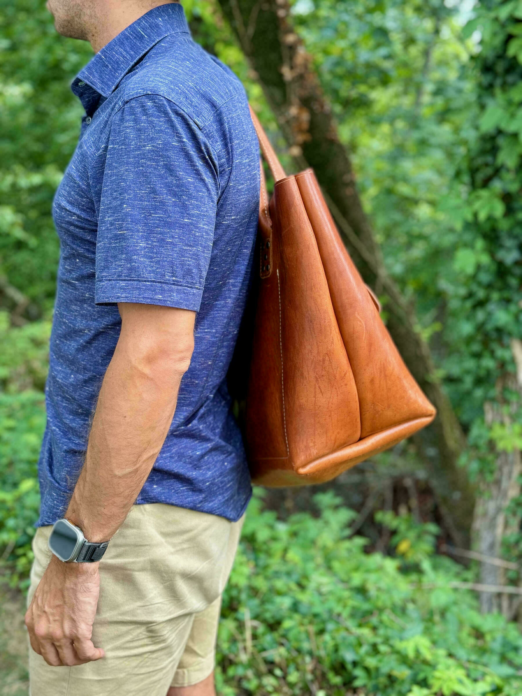 A person wearing a blue shirt and beige shorts is carrying a large brown leather bag over their shoulder while standing in a green outdoor setting.