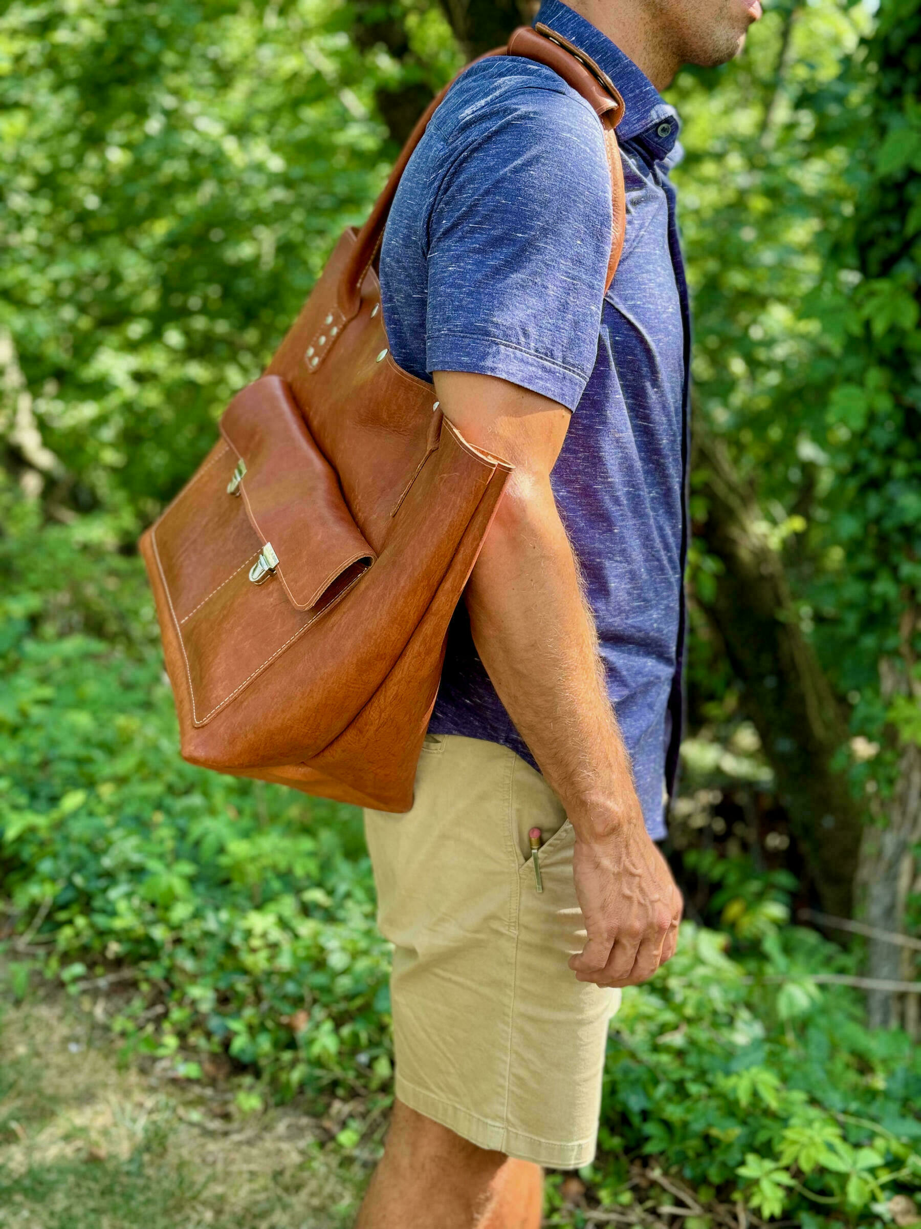 A man, wearing a blue shirt and beige shorts, carries a large brown leather bag over his shoulder in an outdoor setting.