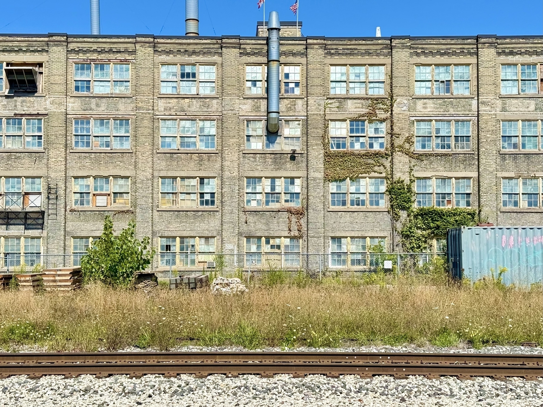 An old industrial building with multiple rows of large windows, many of which are cracked or broken. The structure is partially covered with climbing vines. In front of the building, there is a patch of overgrown grass and weeds, a few small trees, and stacks of wooden pallets. A railroad track runs along the foreground, and a blue sky is visible above.