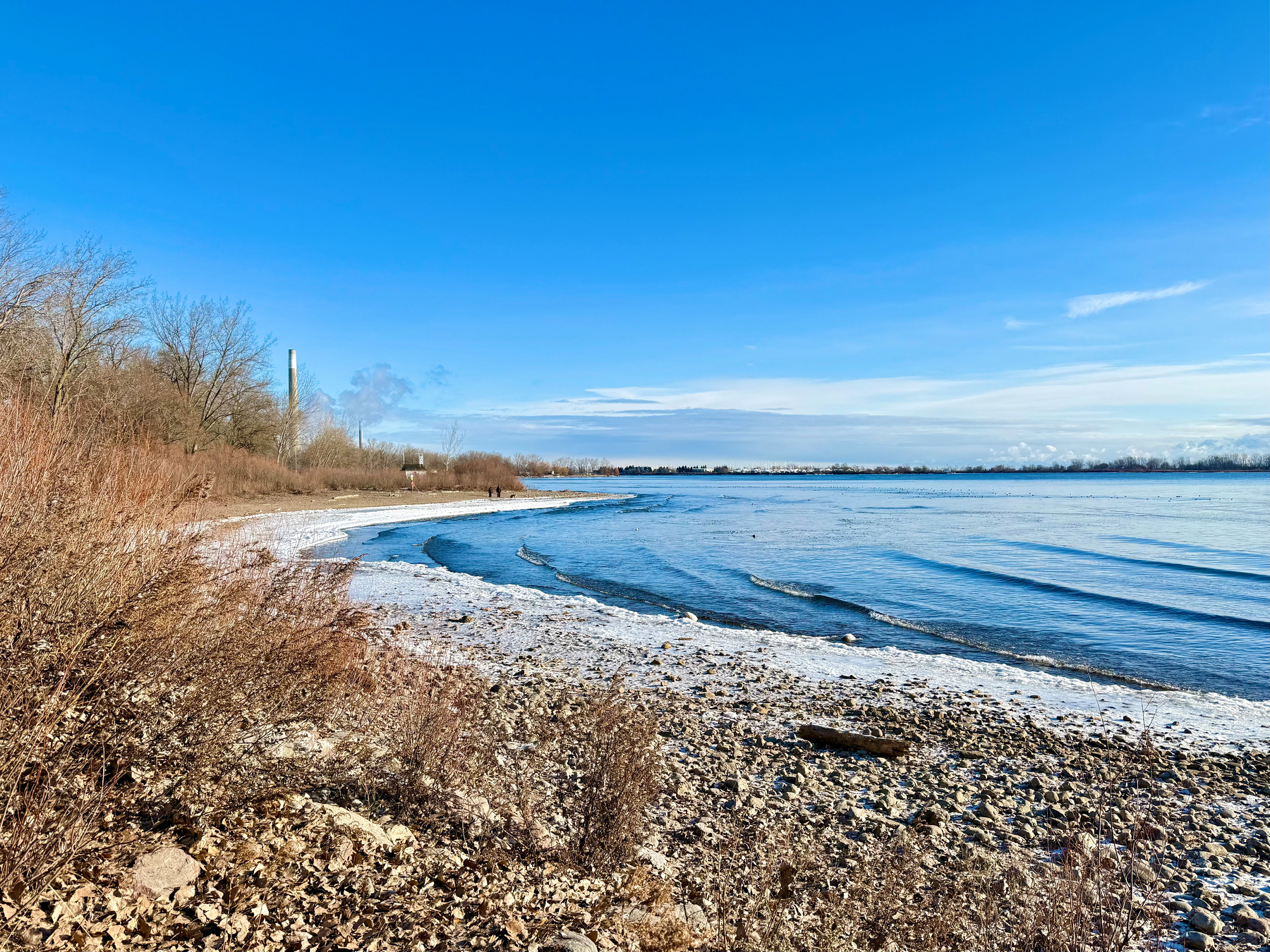 A rocky shoreline with sparse vegetation leads to a calm body of water. The sky is clear and blue, with some clouds on the horizon. In the background, there are leafless trees and a distant smoke stack. The gentle waves lapping at the shore suggest a mild wind. Two people can be seen walking along the beach in the distance.