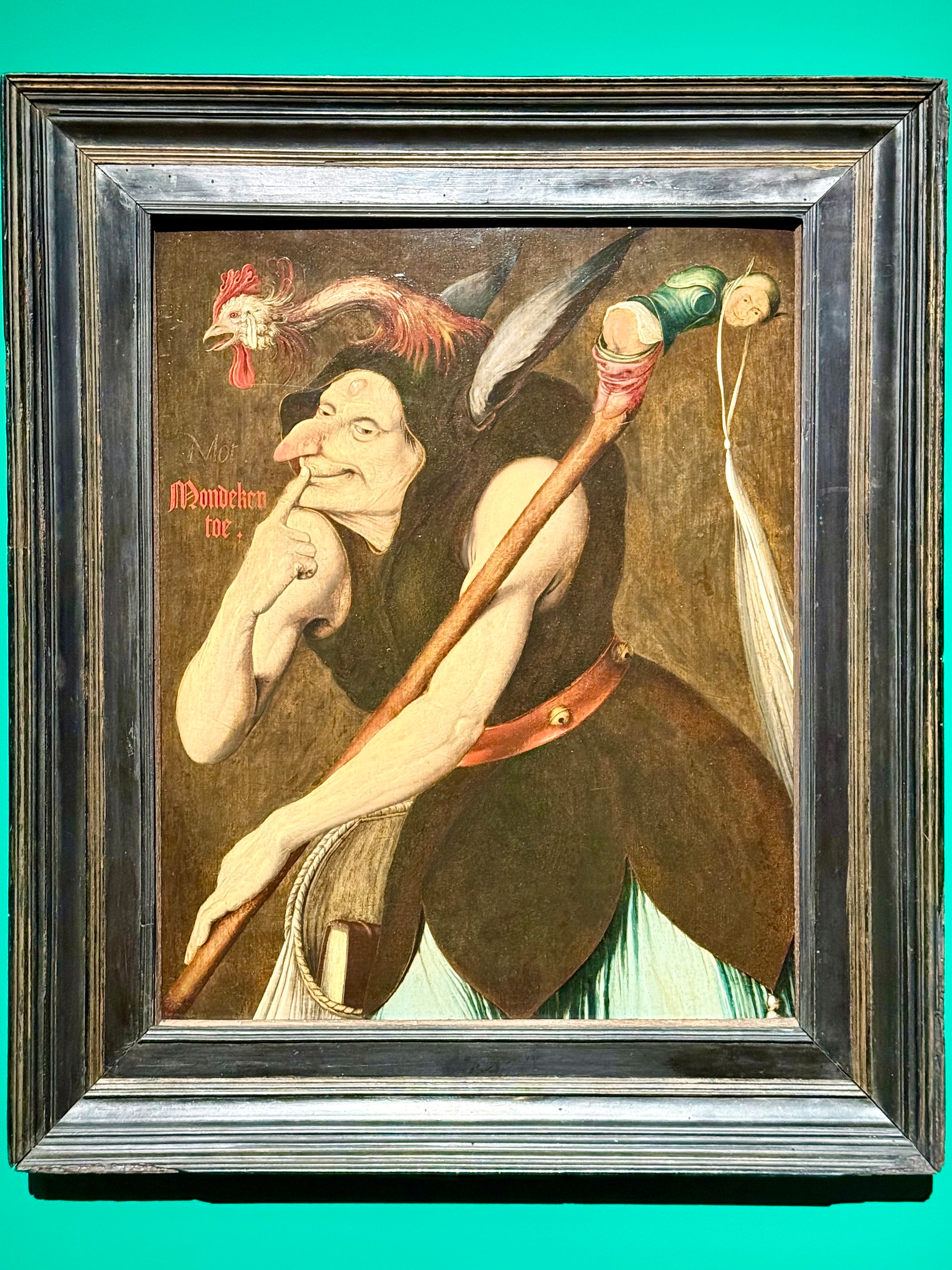 A painting in a dark frame features a surreal figure with an elongated face and a long, pointed nose. The figure holds a walking stick and wears a dark garment with feather-like details. A rooster's head is attached to the figure's headdress, and a human-like face appears on the staff. The background is a muted brown, with stylized writing on the left side.