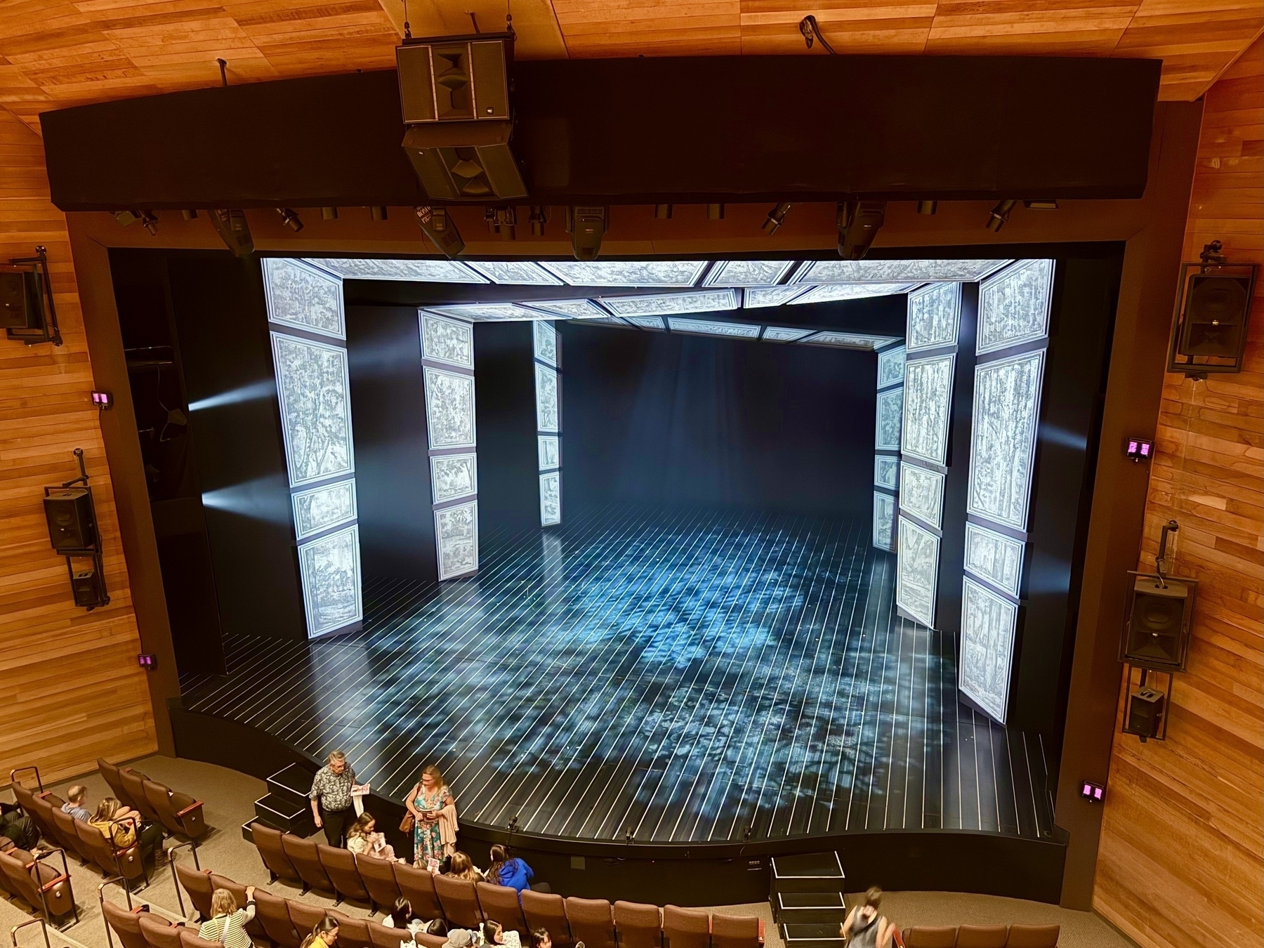 A theater stage is set up with a modern design. The stage features digital panels in a geometric arrangement with black and white patterns resembling architectural drawings. A grid pattern is projected onto the stage floor. The auditorium has wooden walls and rows of empty brown seats, with a few people scattered, conversing or finding their seats. The atmosphere suggests preparation for a performance.