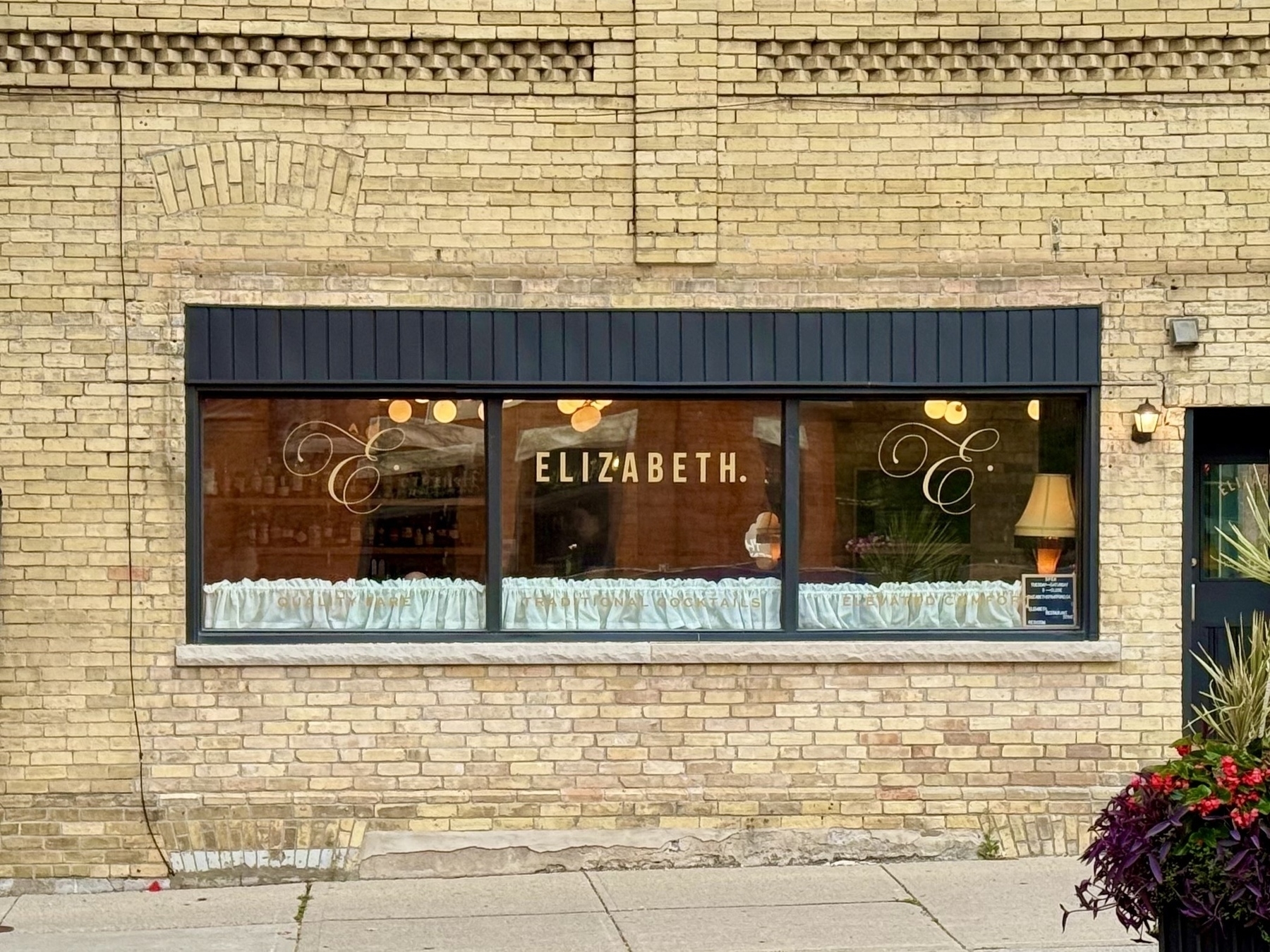 Street view of a brick building with a large rectangular window displaying the word "ELIZABETH." The window is framed by black trim and features decorative swirls on either side of the text. Inside, a lamp and plants are visible. There is a planter with red flowers and purple leaves on the sidewalk in the foreground.