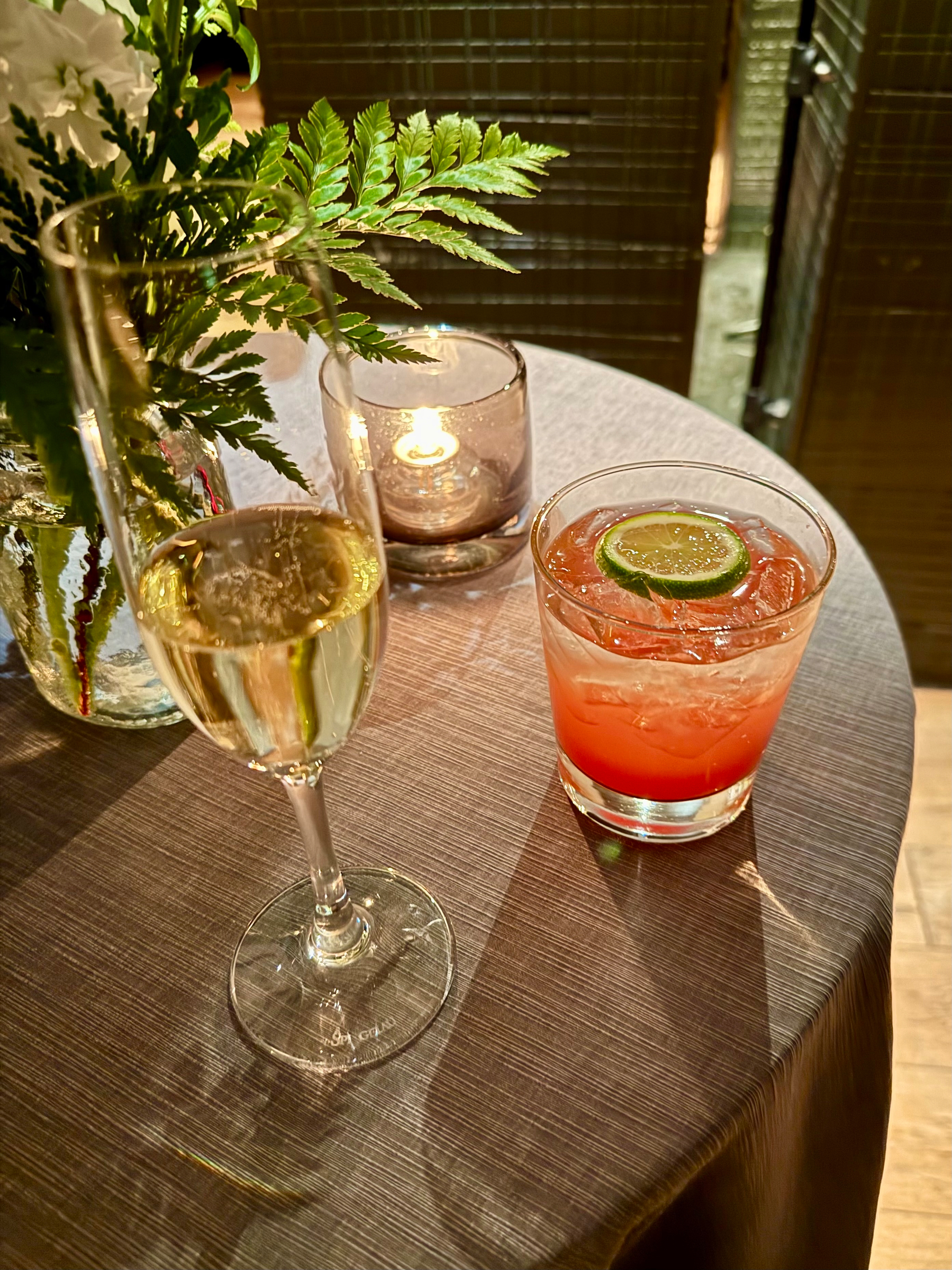 A table setting with a glass of champagne on the left and a cocktail on the right, garnished with a lime slice. A small lit candle in a glass holder is situated between them. Behind the drinks, there is a floral arrangement with green foliage in a vase. The tablecloth is a textured dark fabric, and the scene exudes a warm, inviting ambiance.