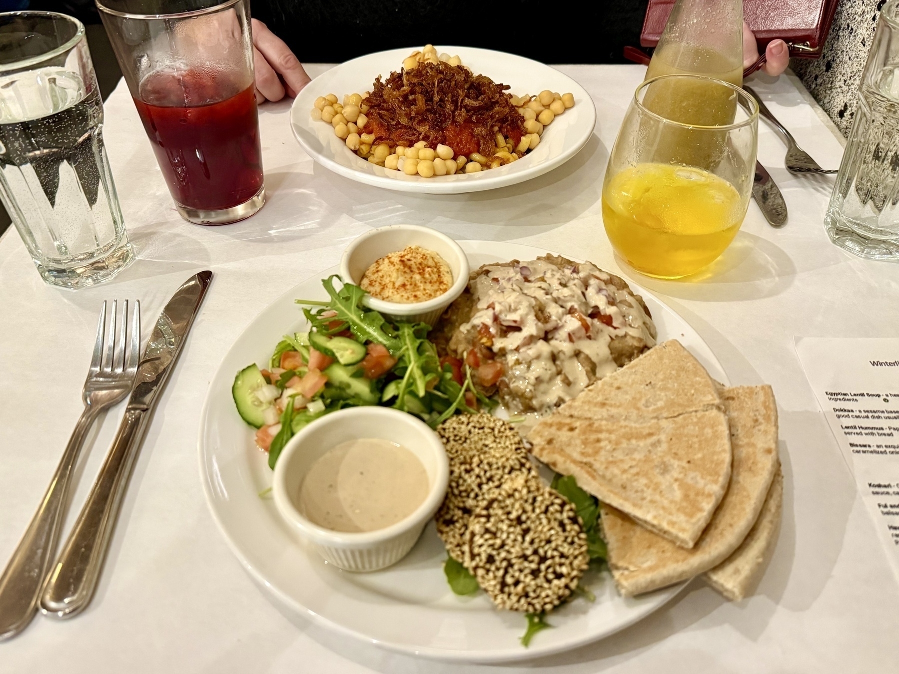 A table set with two plates of Egyptian cuisine in a restaurant. The plate in the foreground contains a variety of items, including a salad with cucumbers, tomatoes, and arugula, a small dish of hummus sprinkled with paprika, a serving of baba ganoush or another eggplant-based dish topped with chopped onions and sauce, sesame-coated falafel, slices of pita bread, and a small bowl of tahini sauce. In the background, another plate holds a dish with chickpeas, a red sauce, and crispy fried onions.