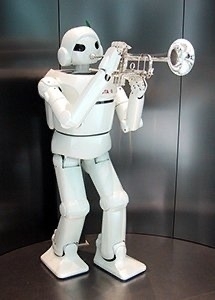 A humanoid robot with a white body, black joints, and a small green detail on its head is playing a silver trumpet. The robot has a futuristic design with a smooth, rounded body and black, visor-like eyes. It is standing in an indoor setting with a metallic background.