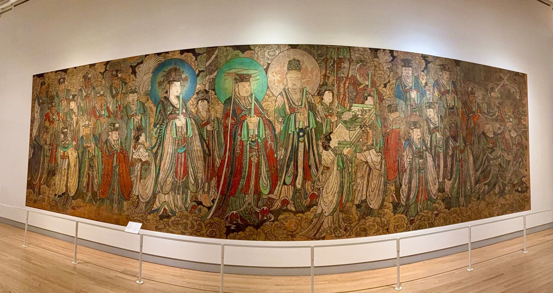 A large, colorful mural of ancient Chinese figures, likely deities or royals, dressed in elaborate, flowing robes with intricate designs. Some figures have halos and hold scrolls or objects. The background is filled with clouds and ornate patterns. The mural is mounted on a white wall in a museum setting with a white railing in front for protection.