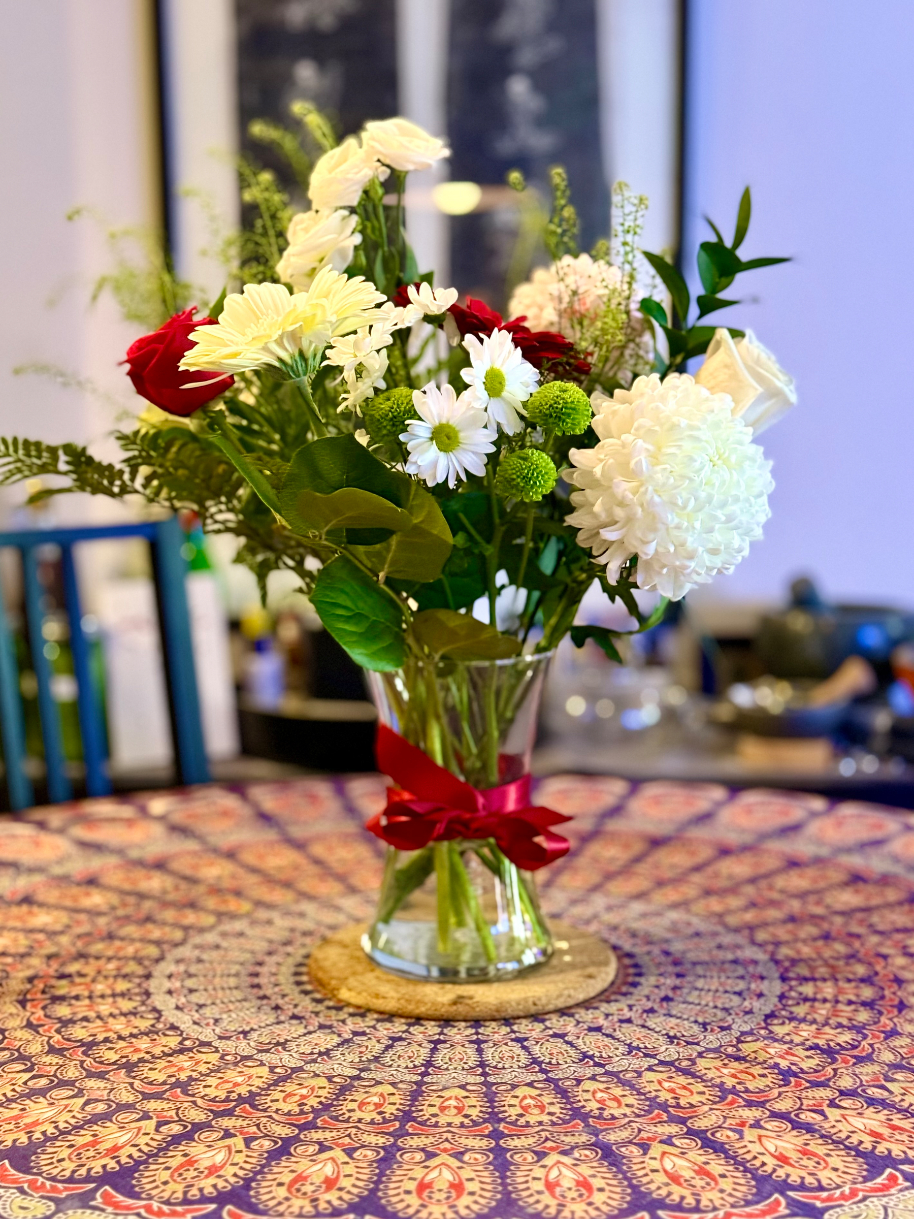 A vase filled with a vibrant bouquet of mixed flowers, including roses and daisies, sits on a patterned tablecloth.