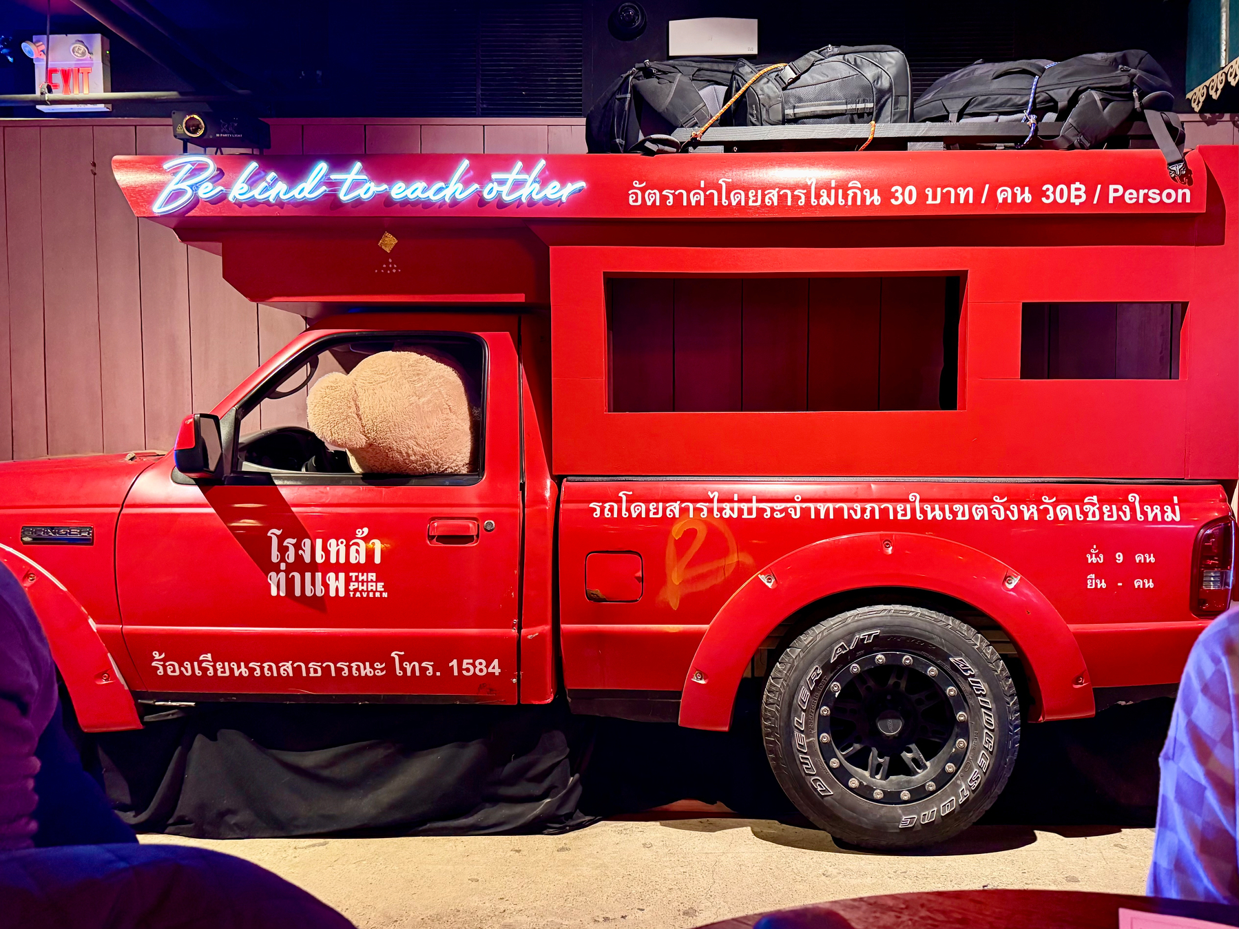 A red vehicle, adorned with luggage on top and text in both English and Thai, including the phrase Be kind to each other, is parked indoors.
