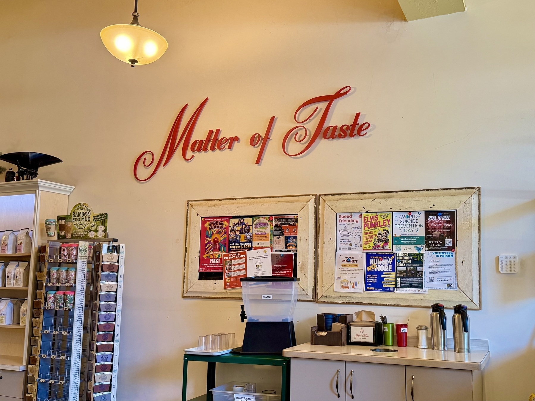 Interior of a cafe with a sign reading "Matter of Taste" in red script on the wall. Below the sign, there's a bulletin board with various colorful posters and flyers. To the left, shelves display products, including boxes and a "Bamboo Eco Mug" sign. There's a counter with coffee containers, cups, and a tissue dispenser. A ceiling light fixture hangs above.