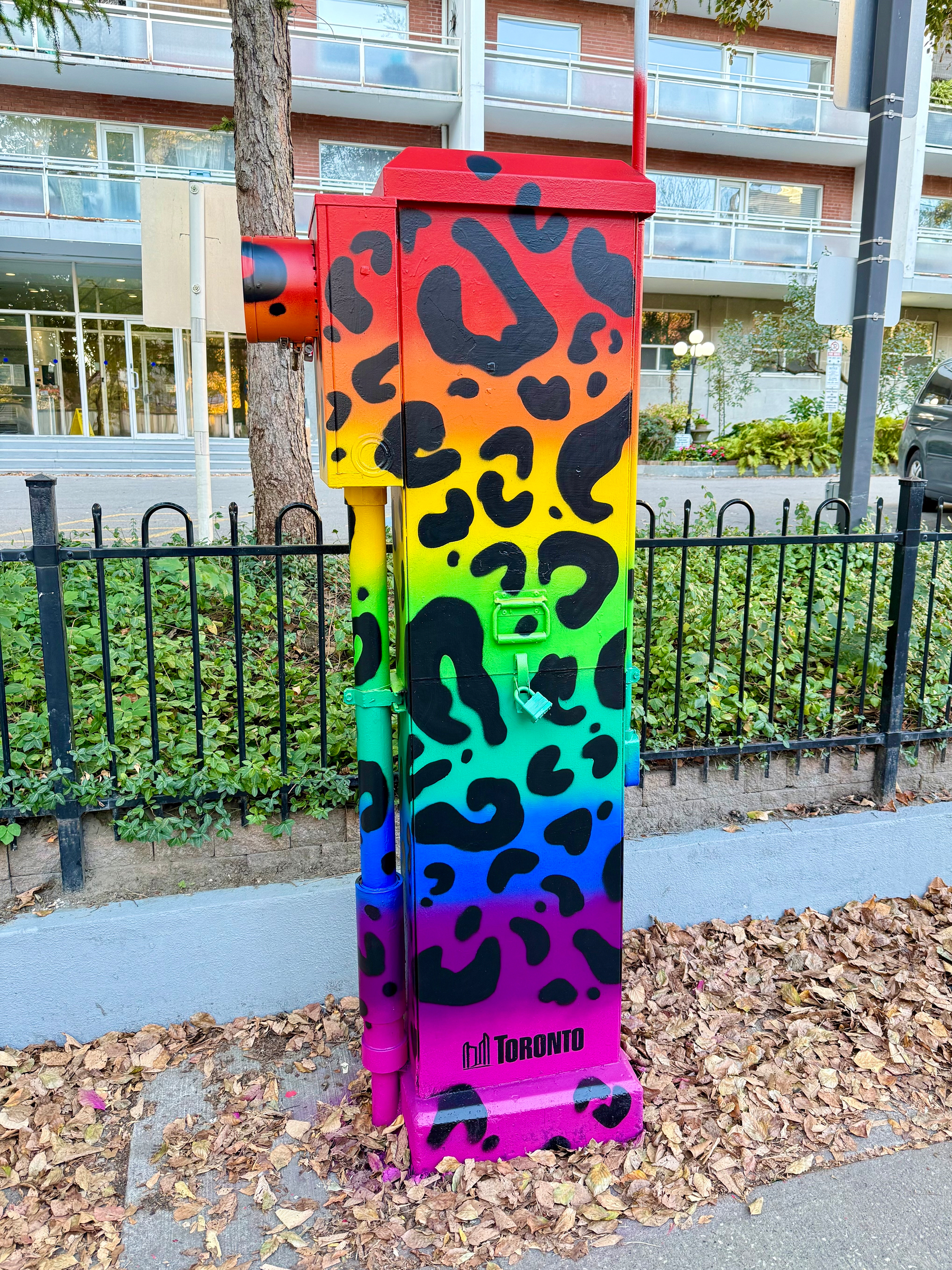 A traffic control box is painted with a vibrant, colorful gradient design, transitioning from red at the top through yellow, green, blue, and ending in pink at the bottom. The surface features black leopard-style spots. The word "Toronto" with a city skyline logo is visible at the bottom. The box is located on a sidewalk covered with fallen leaves, next to a metal fence and a tree, with a building in the background.