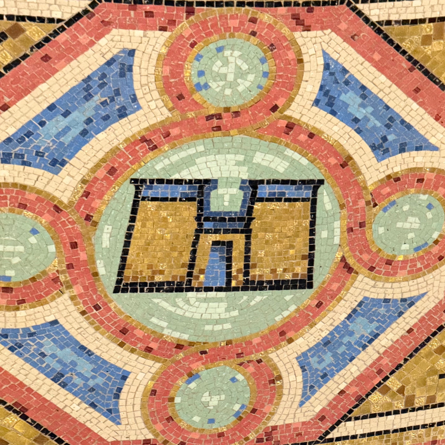 A photo of an intricate mosaic floor featuring the Gates of Nineveh in the center. The design consists of geometric patterns in shades of gold, blue, red, and green, with circular and diamond shapes creating a symmetrical and colorful design surrounding the letter.