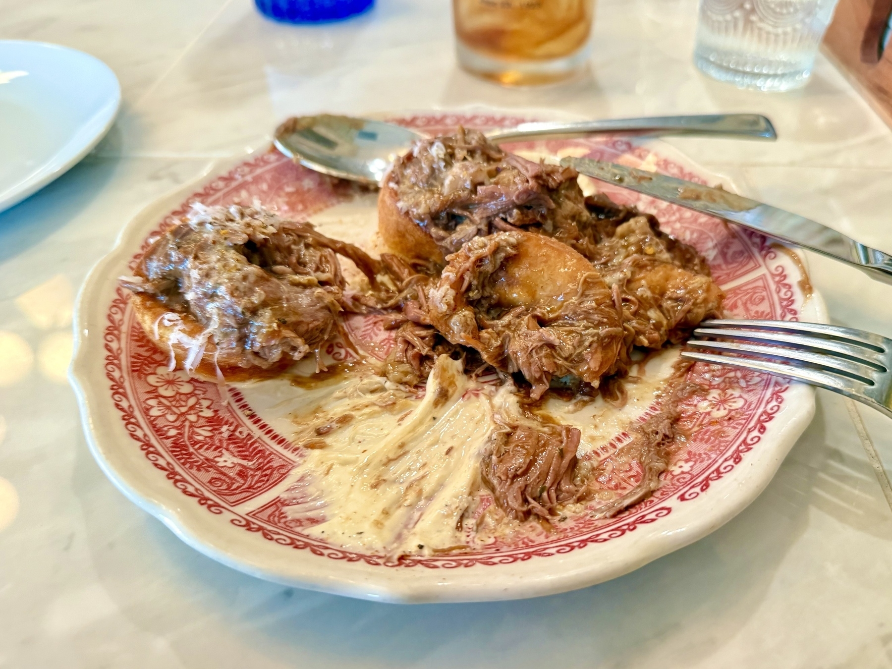 A decorative plate with a red and white floral pattern contains a serving of shredded meat on top of Yorkshire pudding, accompanied by a creamy sauce. A fork and knife are placed on the plate. In the background, there are glassware items including a clear glass and a blue glass, suggesting a meal setting on a marble table.
