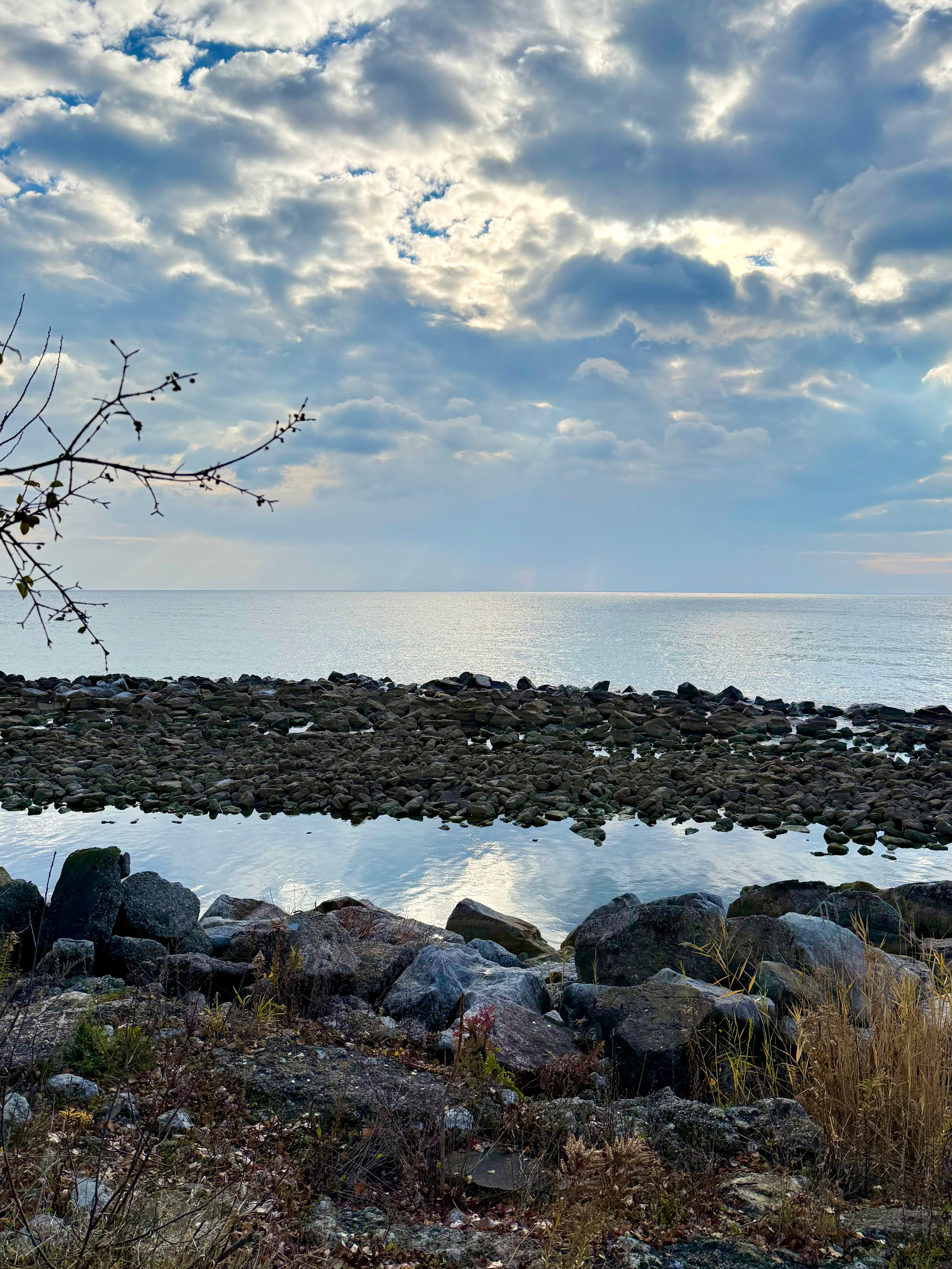 A rocky shoreline leads to a calm sea under a cloudy sky with sunlight peeking through.