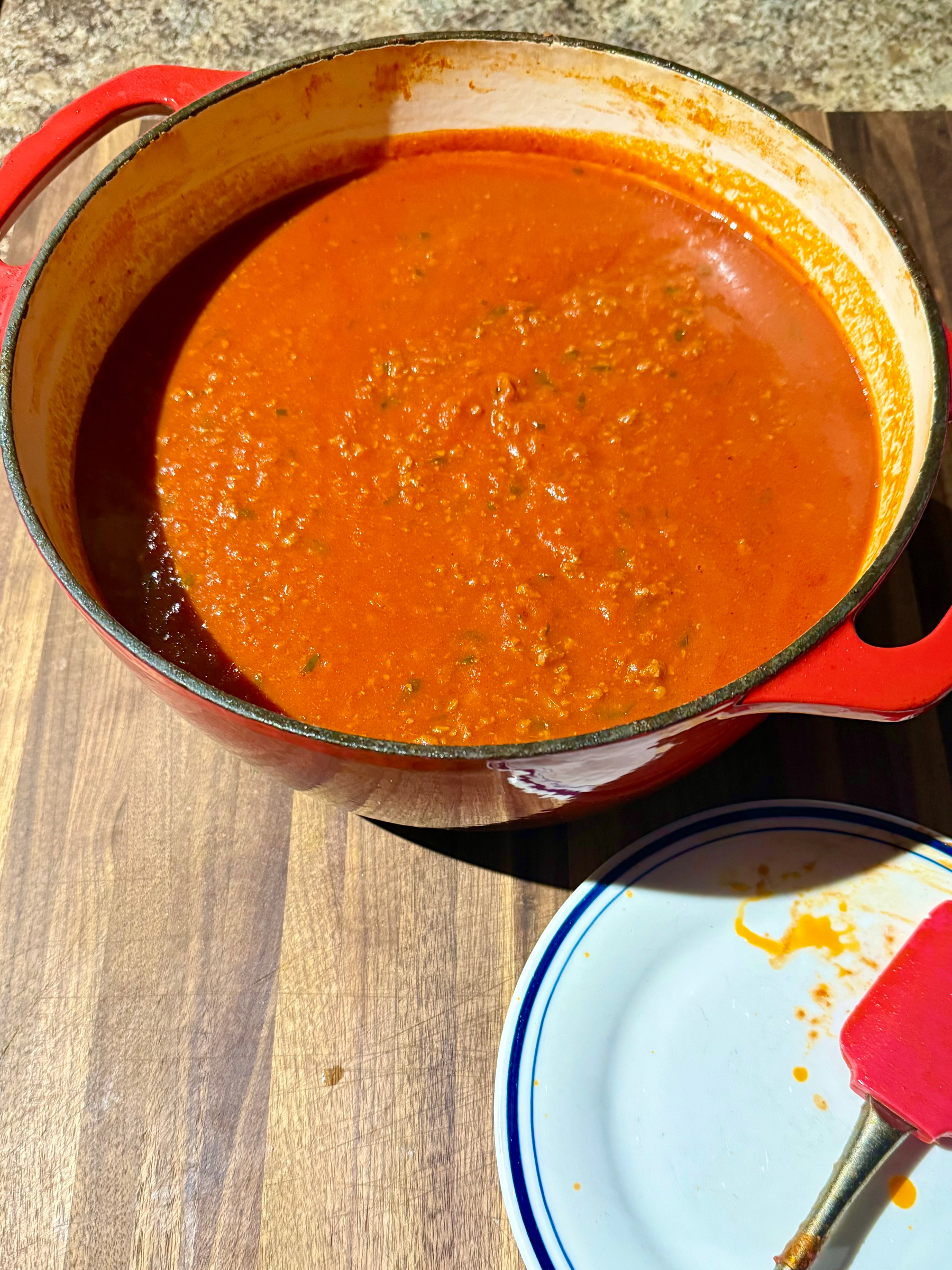 A red pot filled with thick tomato sauce, possibly with herbs and spices, resting on a wooden surface. To the right is a white plate with a blue trim, showing a red spatula with some sauce residue. The setting suggests preparation or cooking in progress.