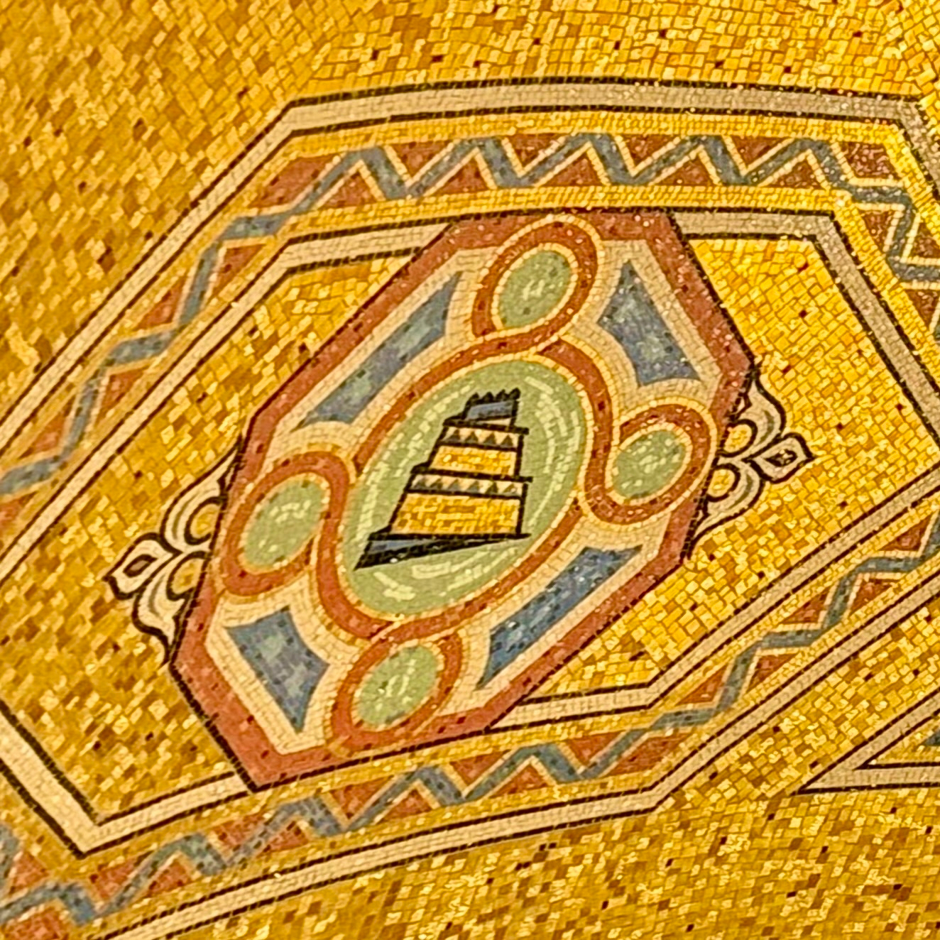 A detailed mosaic with a central emblem of a Babylon surrounded by circular and geometric patterns. The background is primarily gold with decorative elements in red, green, blue, and white, featuring intricate designs and motifs.