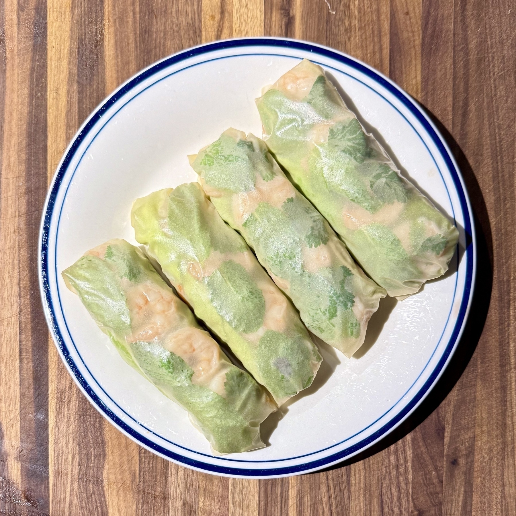 Four fresh rolls wrapped in translucent rice paper on a white plate with a blue rim, set on a wooden surface. The rolls contain visible shrimp, green lettuce, and herbs.
