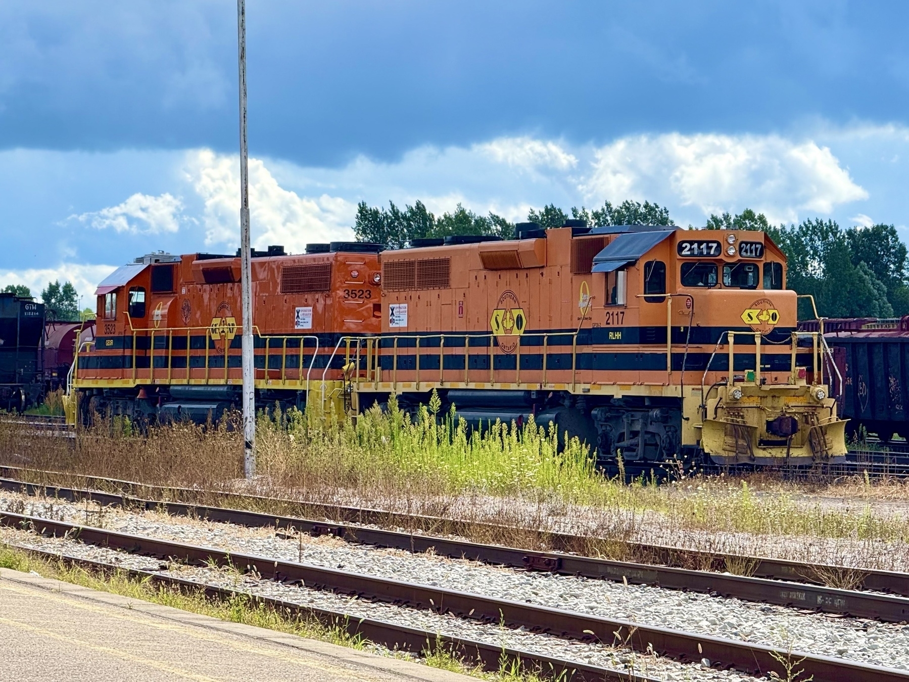 An orange and black locomotive situated on railway tracks with a backdrop of lush green trees and a partly cloudy blue sky. The train features distinct numbering and the SOO Line logo. Tall weeds and grass grow alongside the tracks.